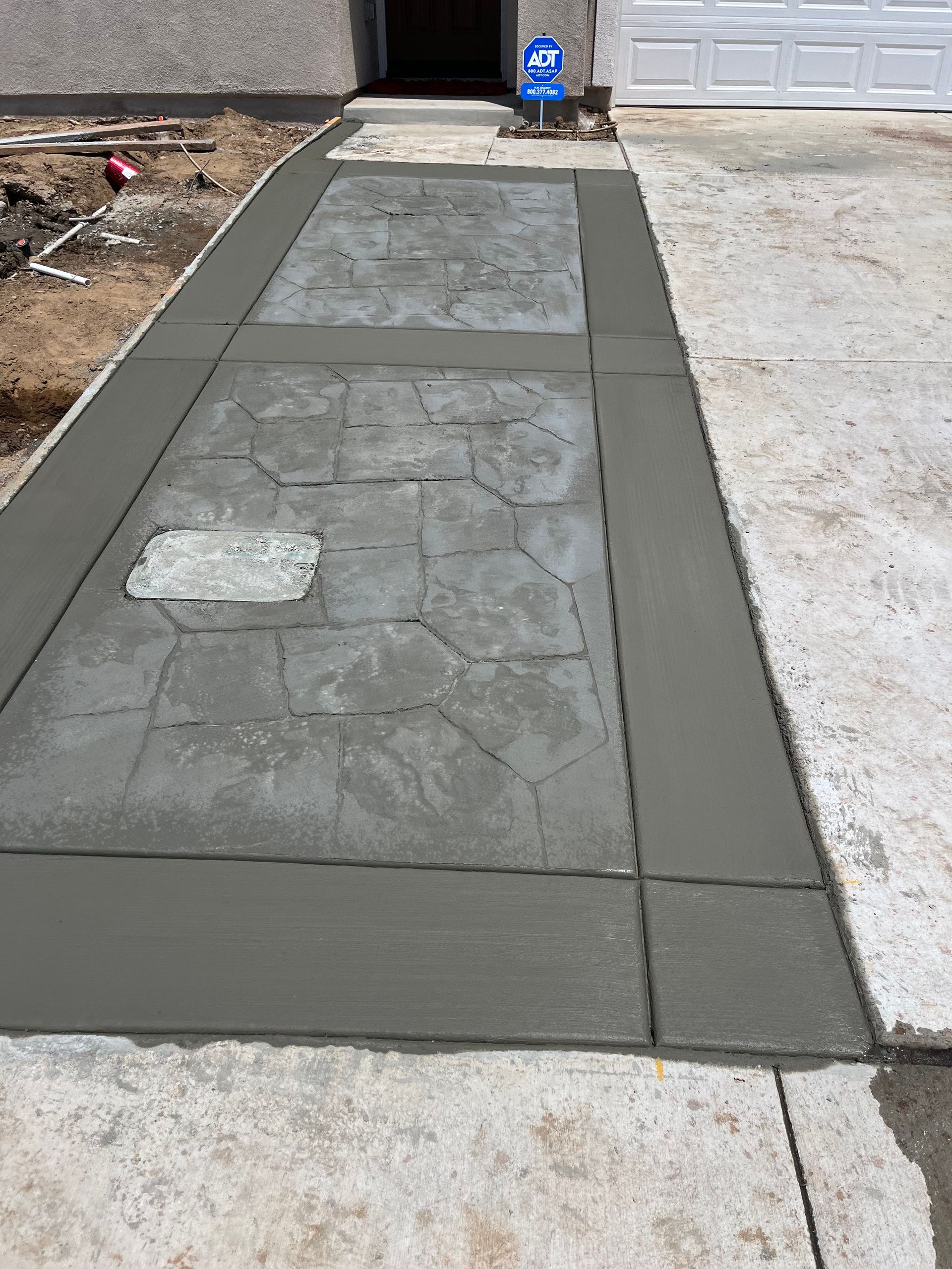 Newly poured concrete sidewalk with stone-patterned walkway, bordered in gray concrete, leads to a front door.