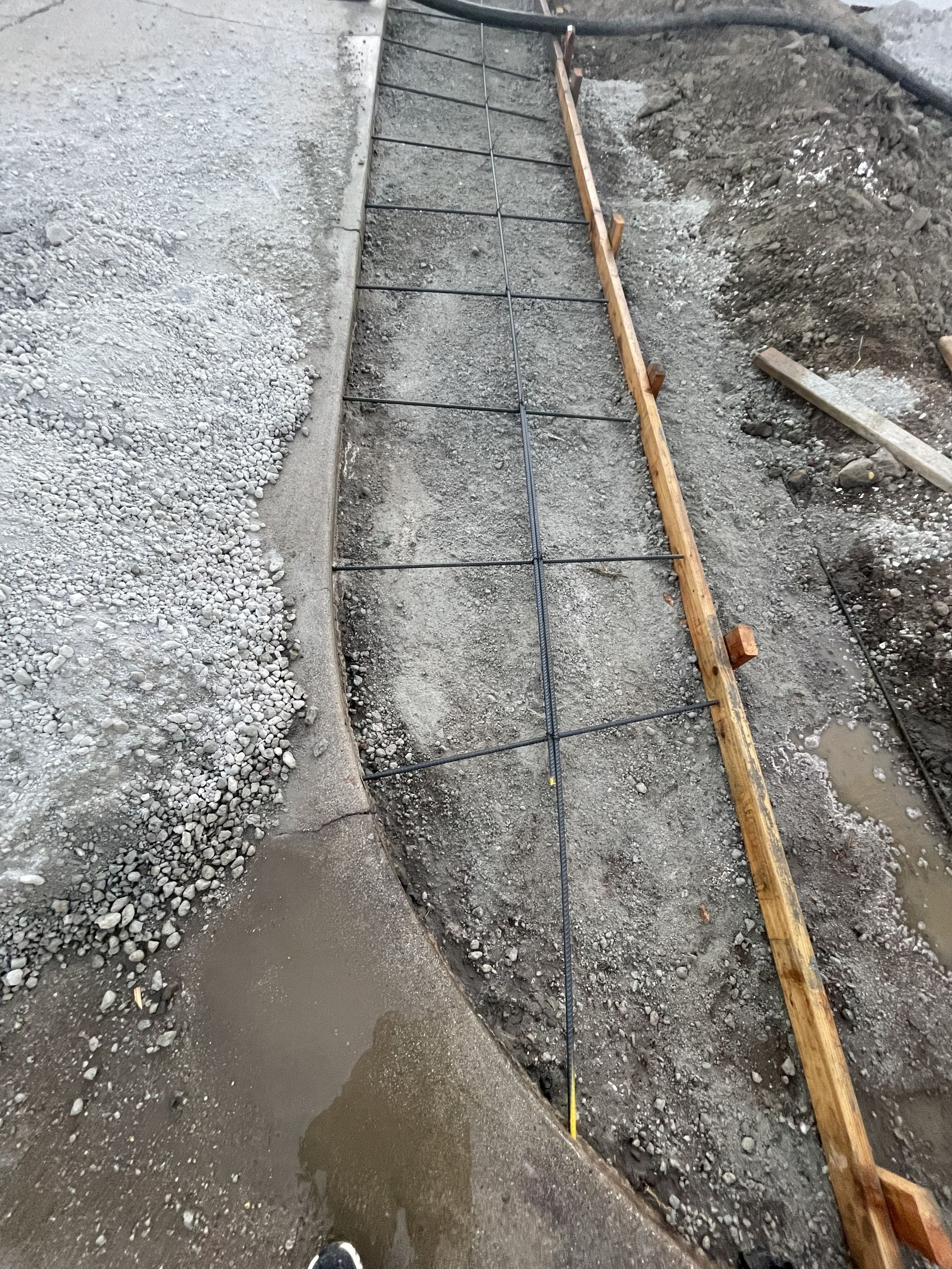Construction site. Concrete path with rebar grid laid, surrounded by gravel and wet pavement.
