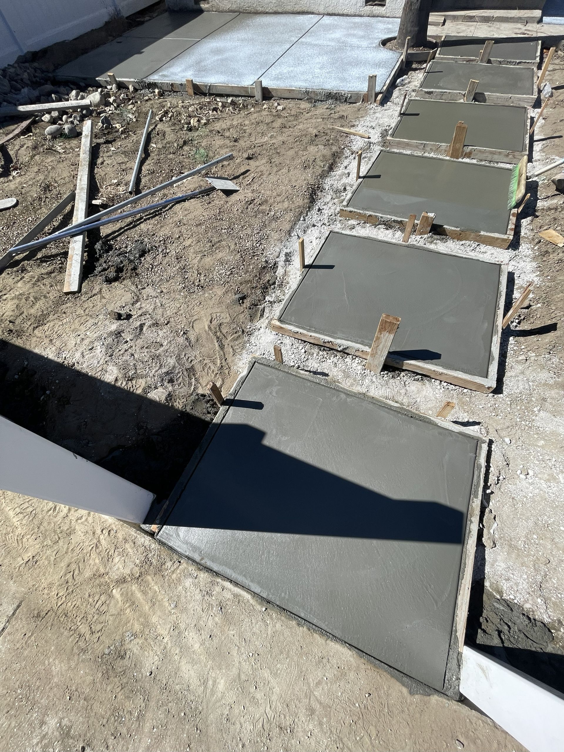 Freshly poured concrete stepping stones, forming a path, sit on the dirt ground with construction materials scattered around.