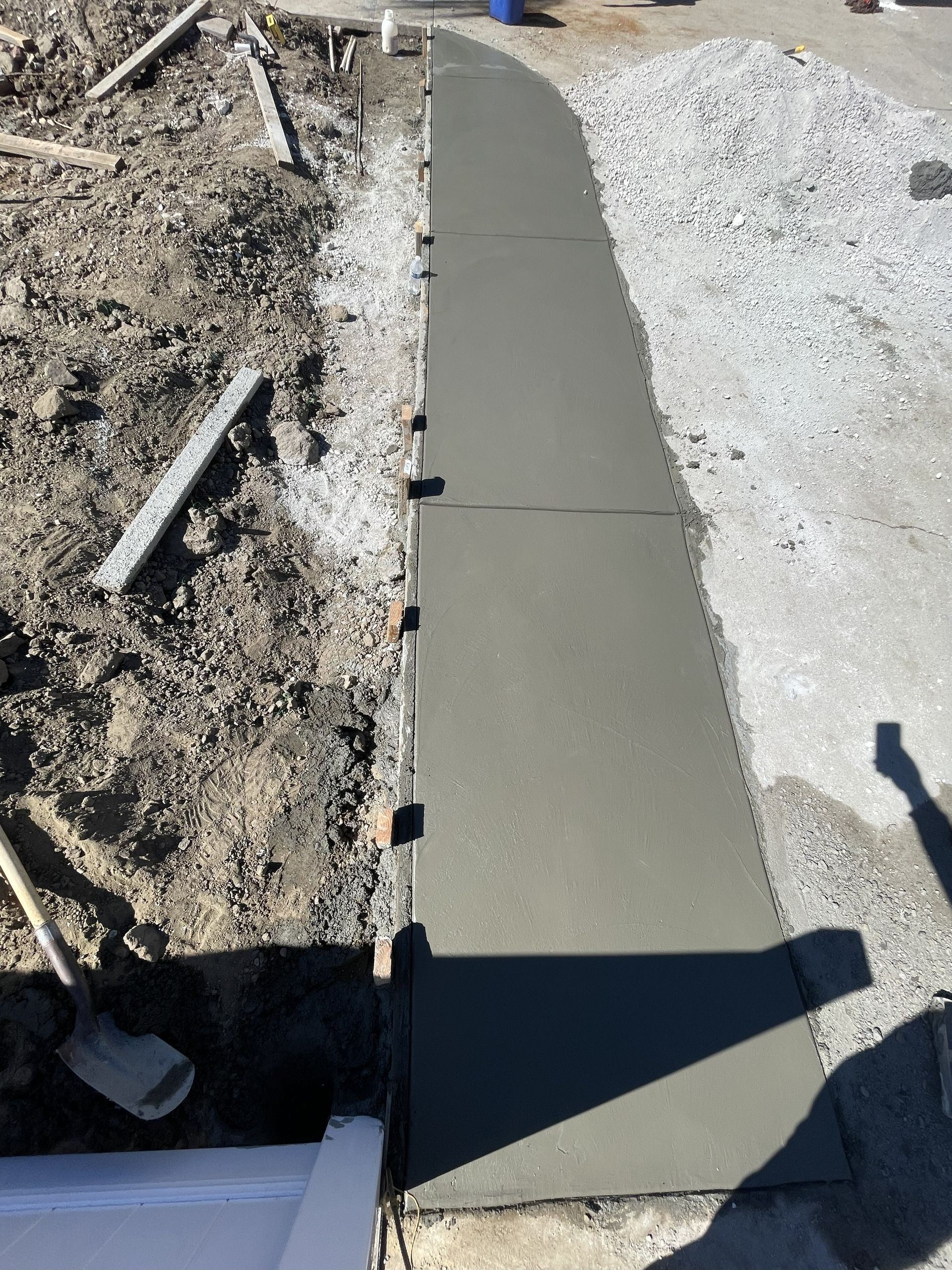 Freshly poured concrete sidewalk next to dirt and gravel, with a shovel and wooden forms visible.