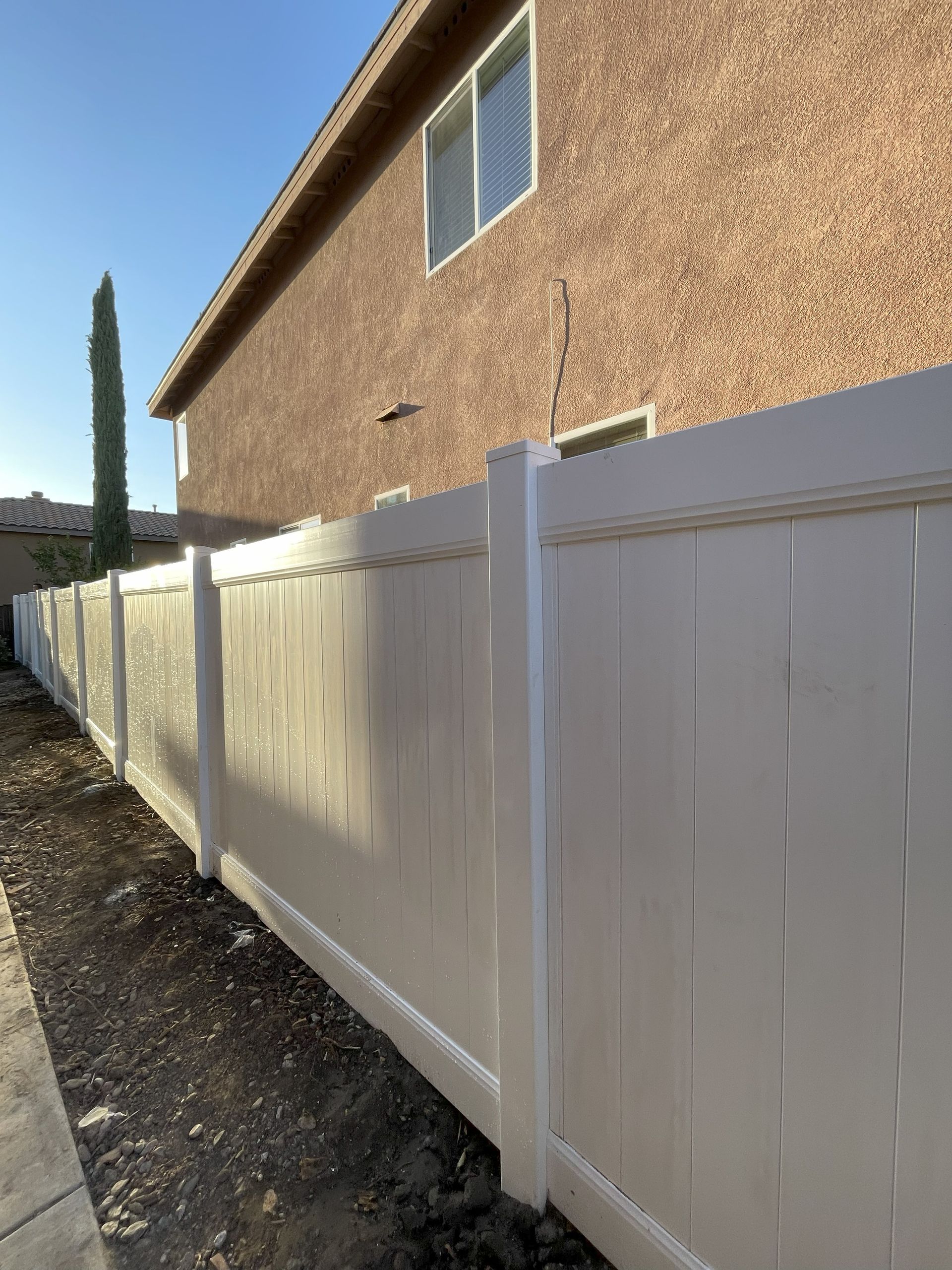 White vinyl fence alongside a building with tan stucco siding under a blue sky.