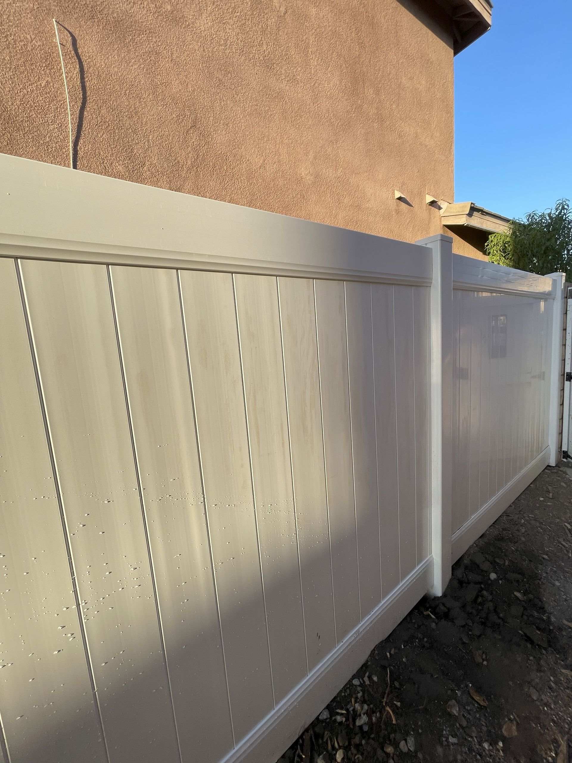 Beige vinyl fence along a textured brown wall, outdoors on a sunny day.