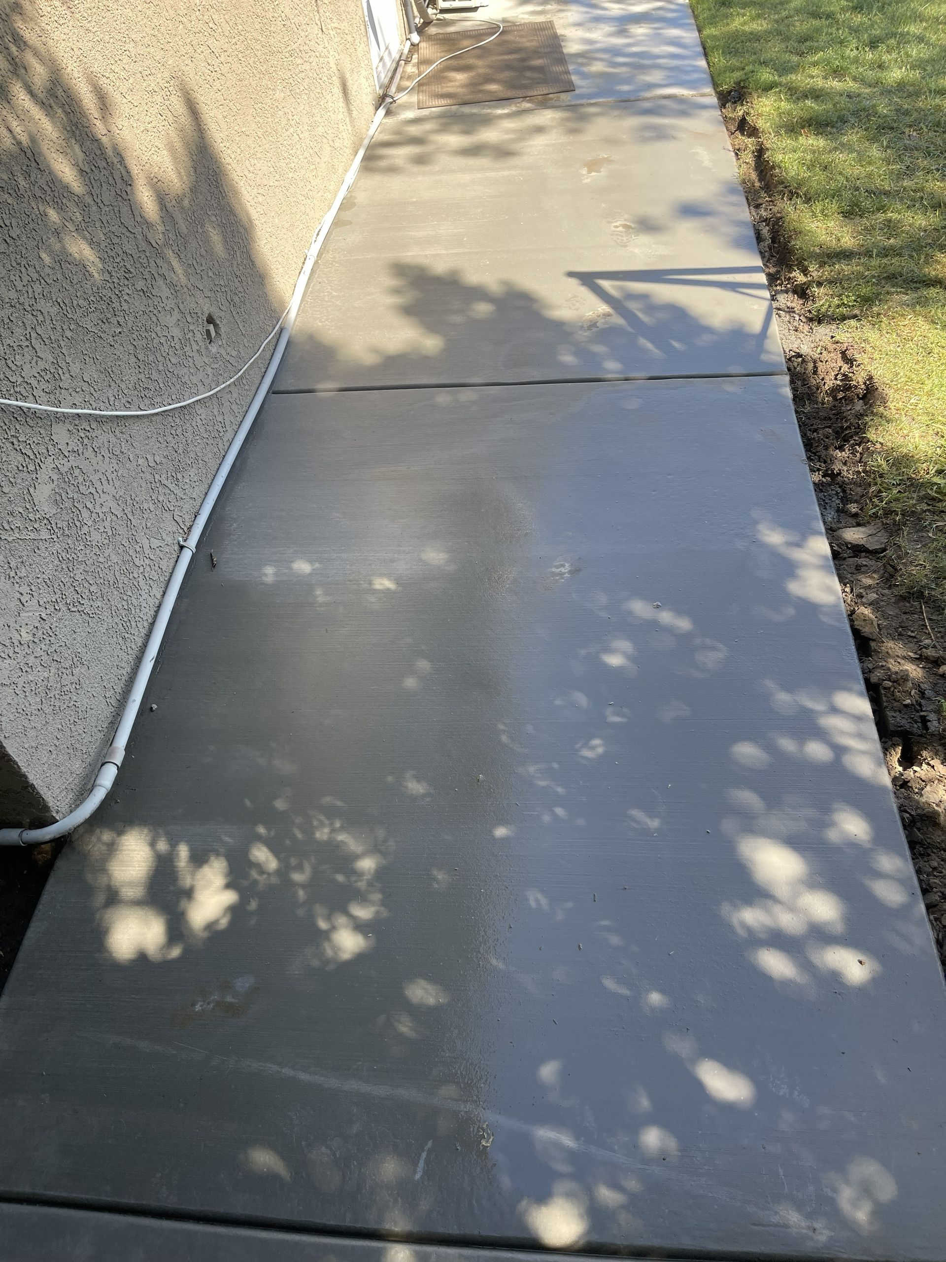 A newly poured concrete walkway next to a light-colored stucco wall and grass.
