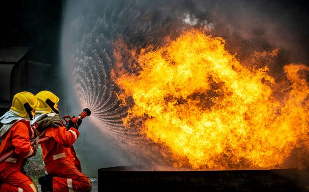 Firefighters in orange suits extinguish a large fire with a water hose.