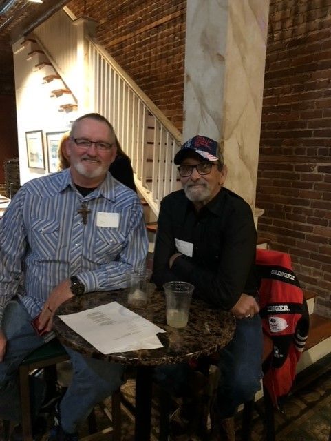 Two men are sitting at a table in front of a brick wall.