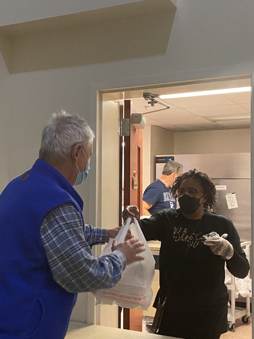 A man in a blue vest is giving a woman a bag of food