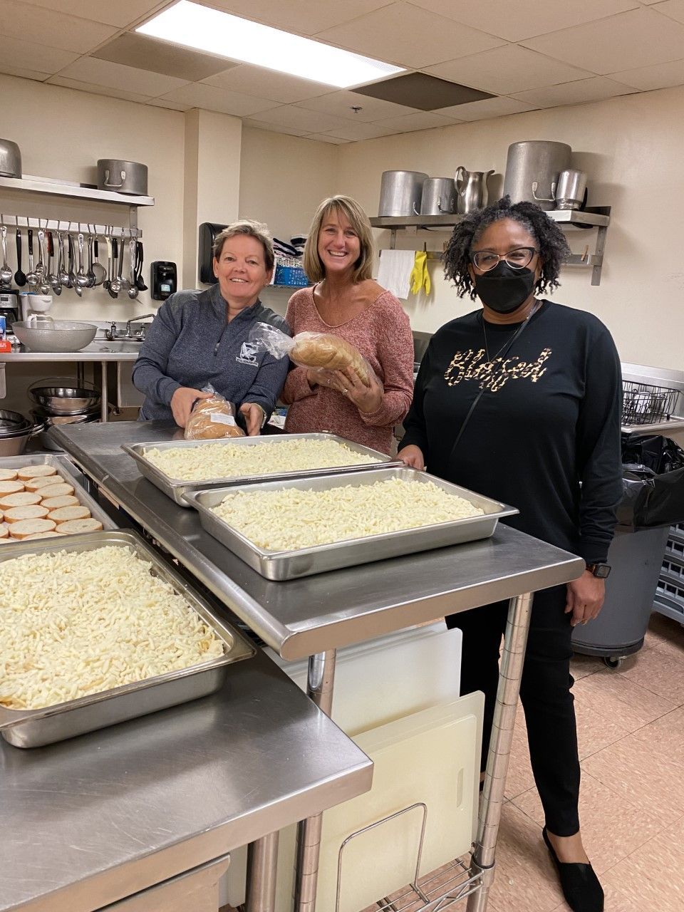 Three women are standing in a kitchen holding trays of food.