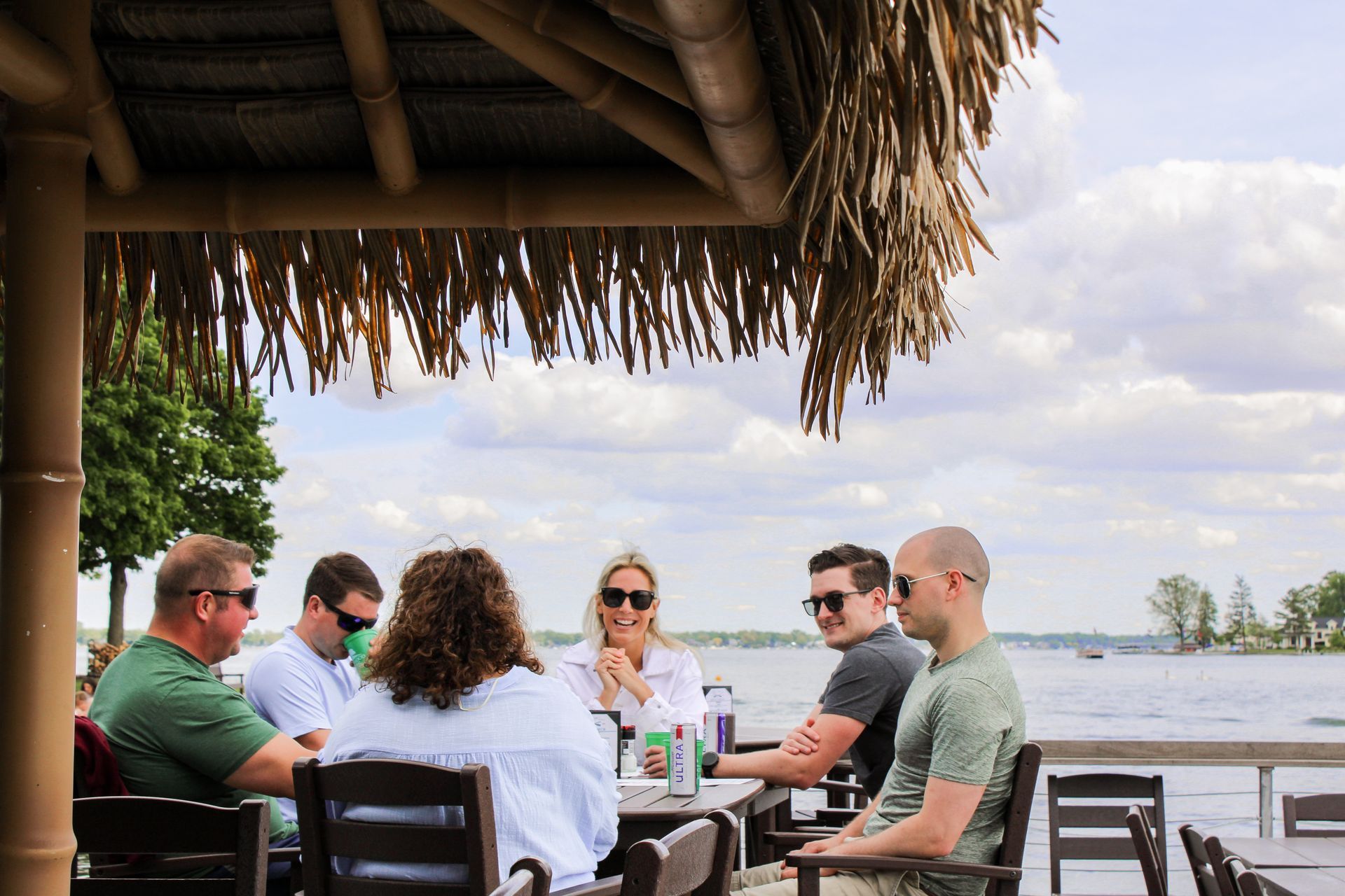 A group of people are sitting at a table under a thatched roof overlooking a body of water.