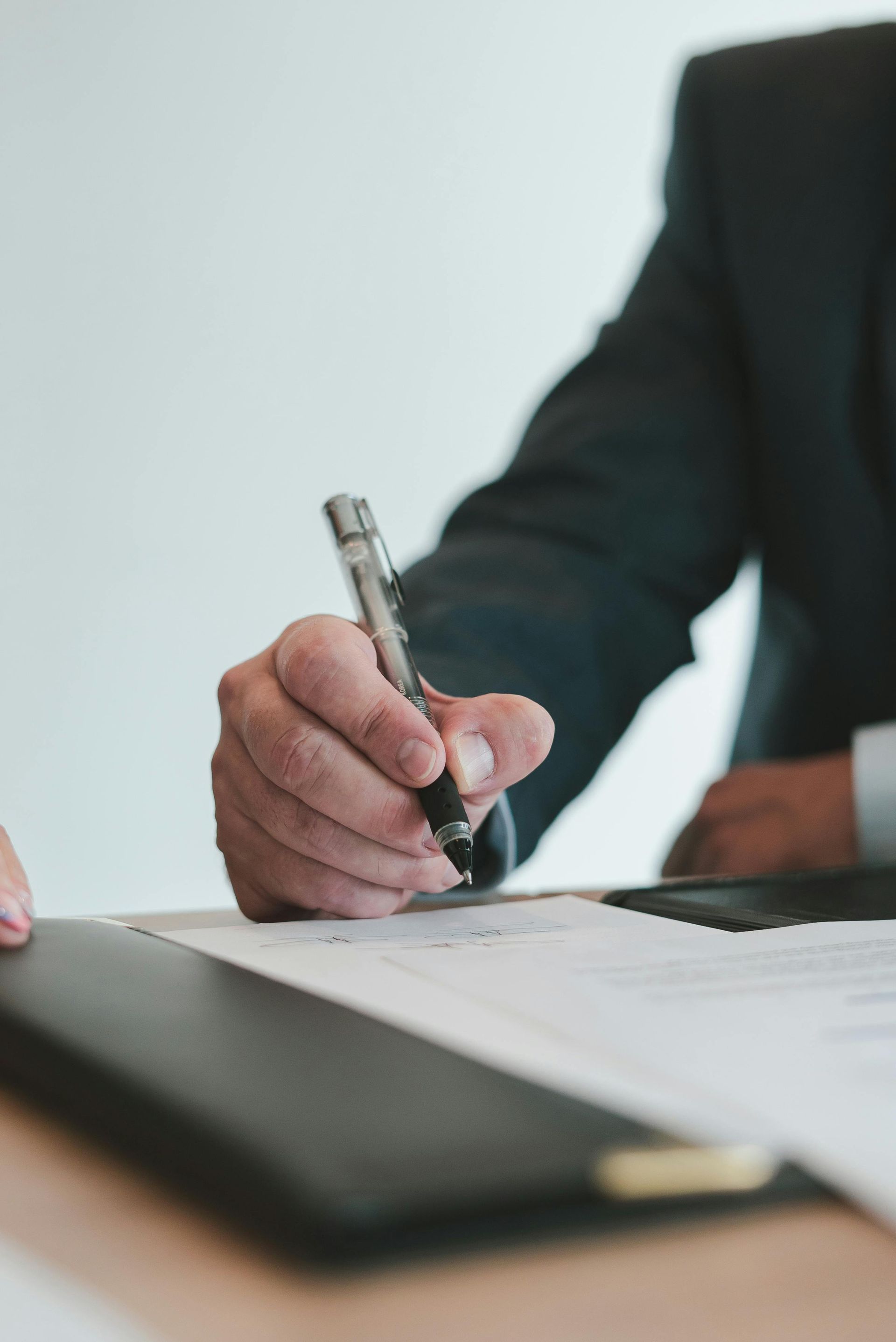 Person in suit signing a document with a pen at a desk.