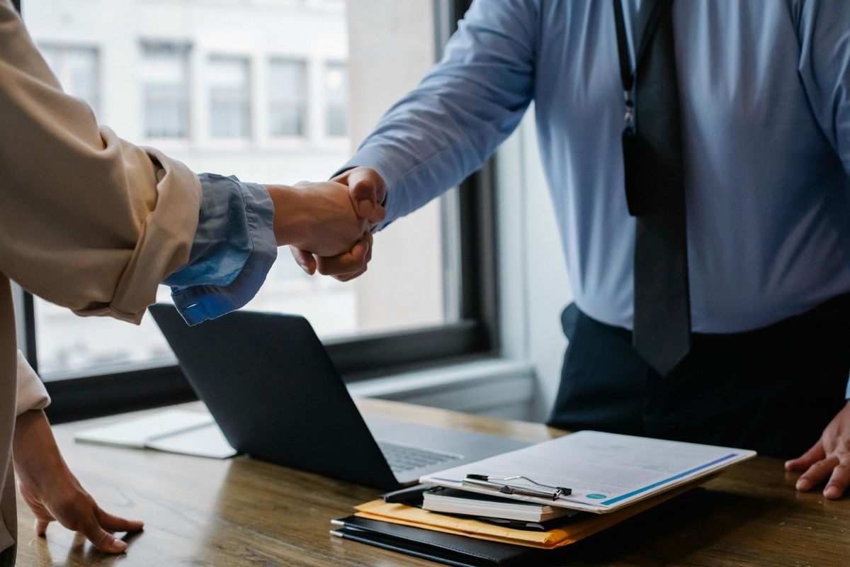 Two people shaking hands over a desk with a laptop and paperwork.