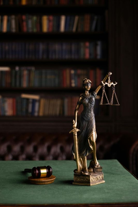 Statue of Lady Justice with scales and sword, gavel on green table, library backdrop.
