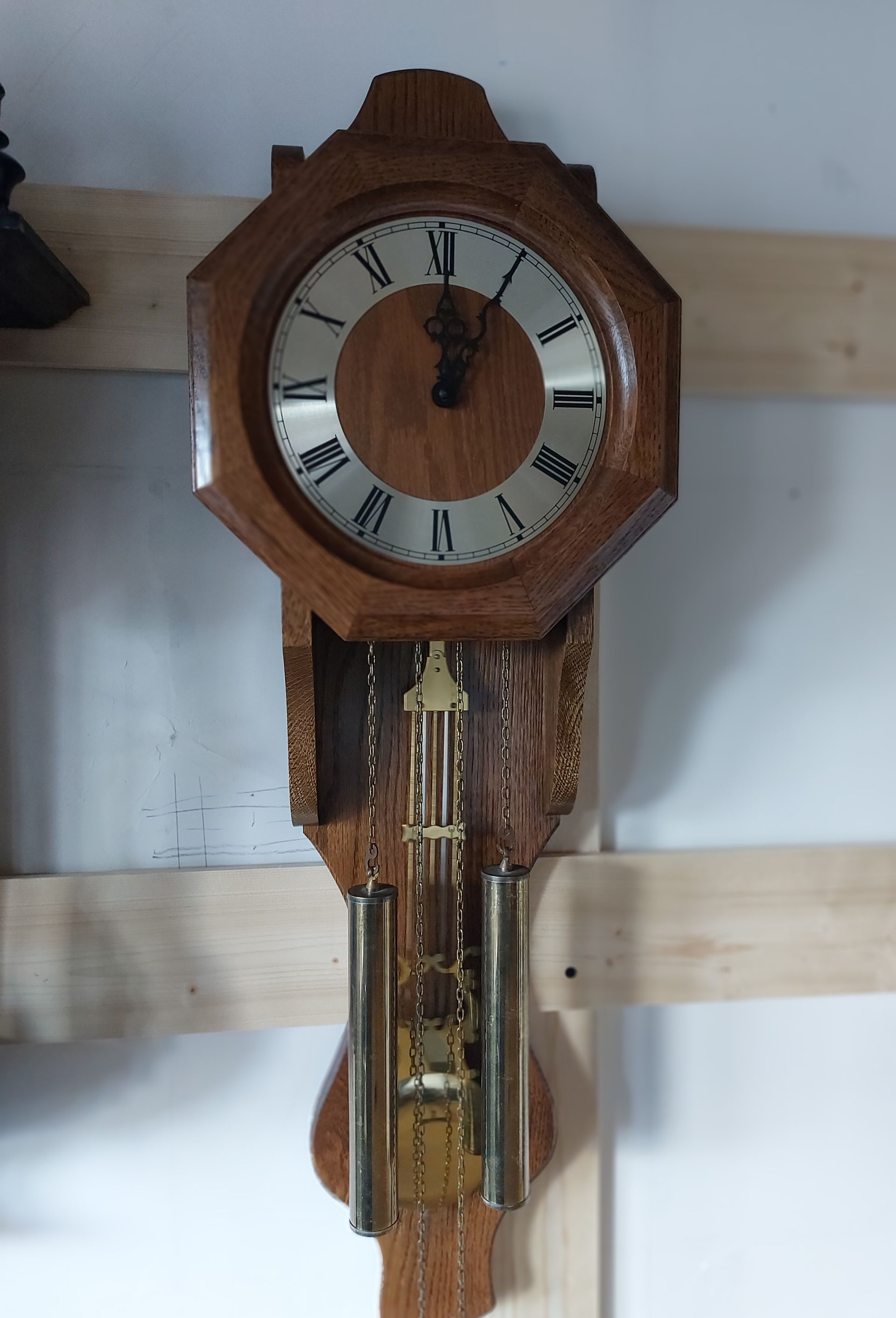 Wandklok van hout met Romeinse cijfers en slinger, gemonteerd op een lichte muur.