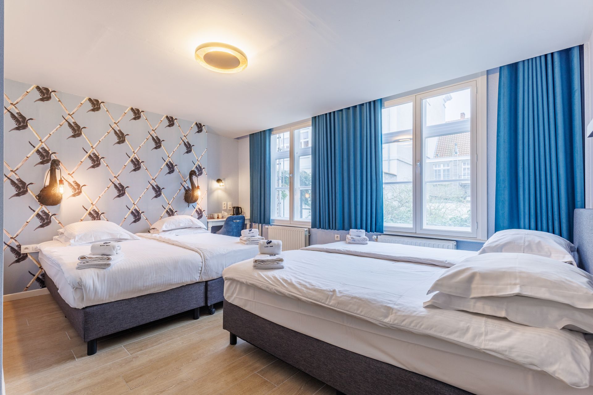 A hotel room with two twin beds featuring white bedding, patterned bird wallpaper, and bright blue floor-length curtains.