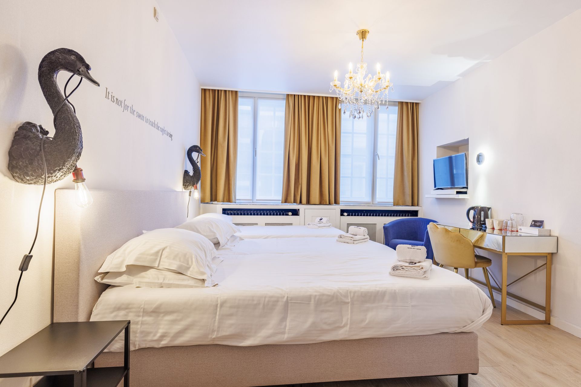 Hotel room featuring a large bed with white linens, two swan wall lamps, gold curtains, and a desk with a blue chair.
