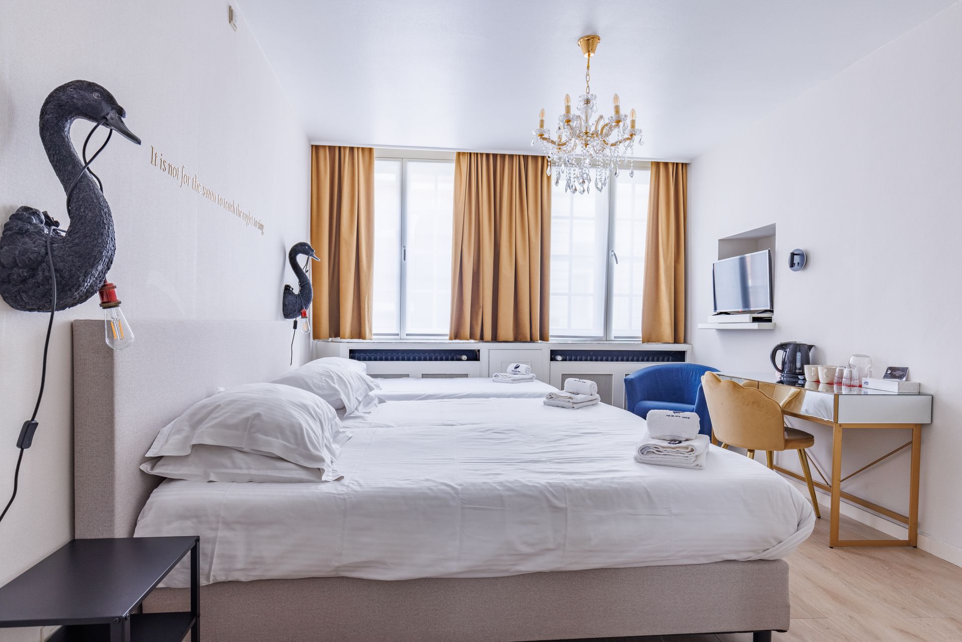 A hotel room featuring two joined beds, gold curtains, a decorative black swan wall light, and a desk with a blue chair.