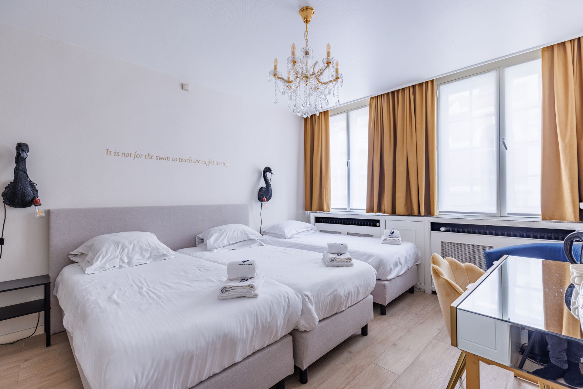 A bright hotel bedroom featuring three white twin beds, gold curtains, and a crystal chandelier.