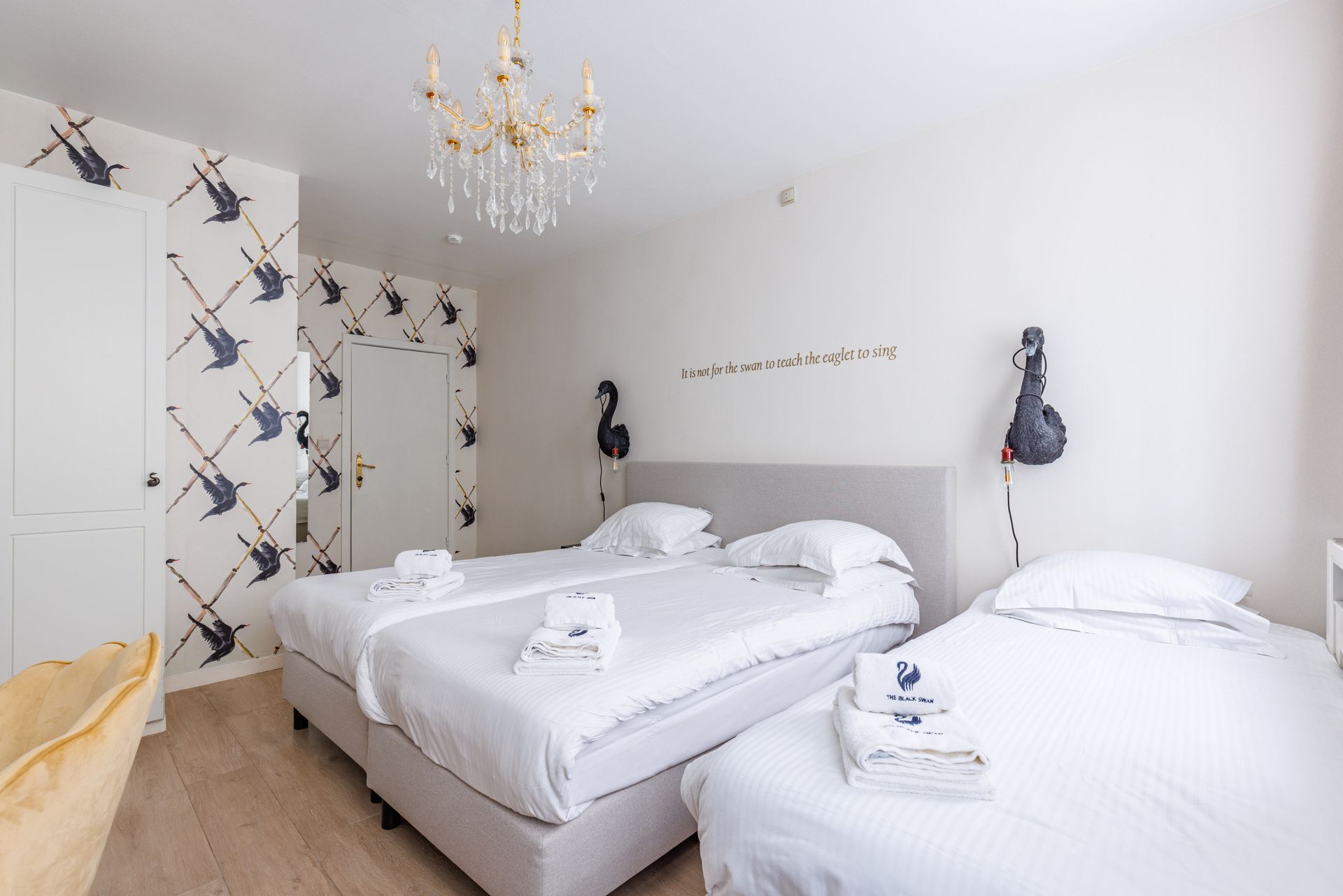 A hotel room with two beds, white bedding, bird-patterned wallpaper, and a crystal chandelier.