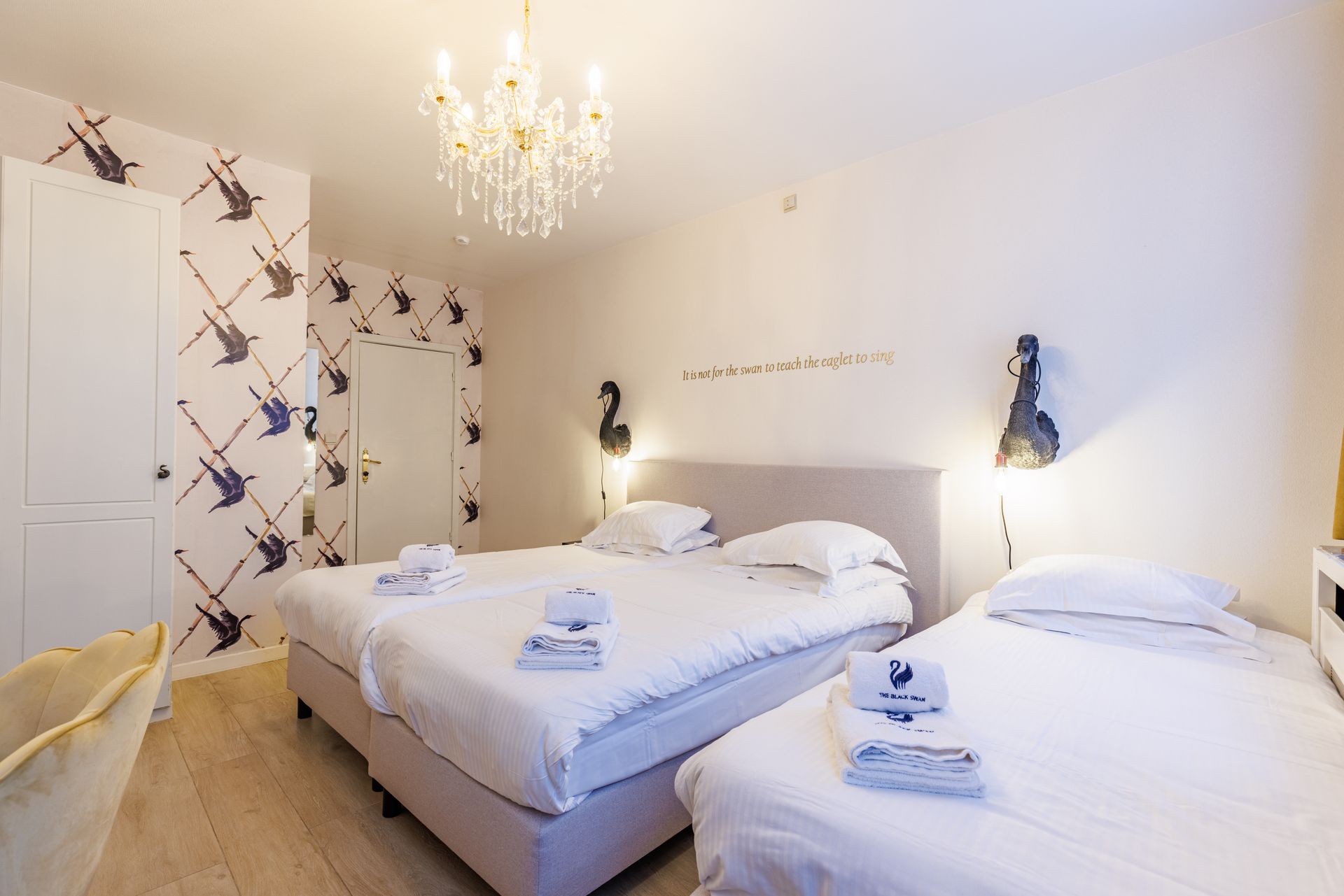 A hotel room with two white beds, bird-patterned wallpaper, a crystal chandelier, and wall-mounted swan light fixtures.