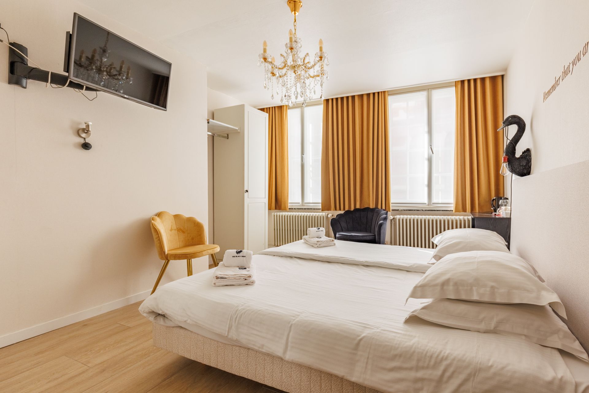 A tidy hotel room with two white beds, yellow curtains, a matching yellow chair, and a flat-screen TV on the wall.