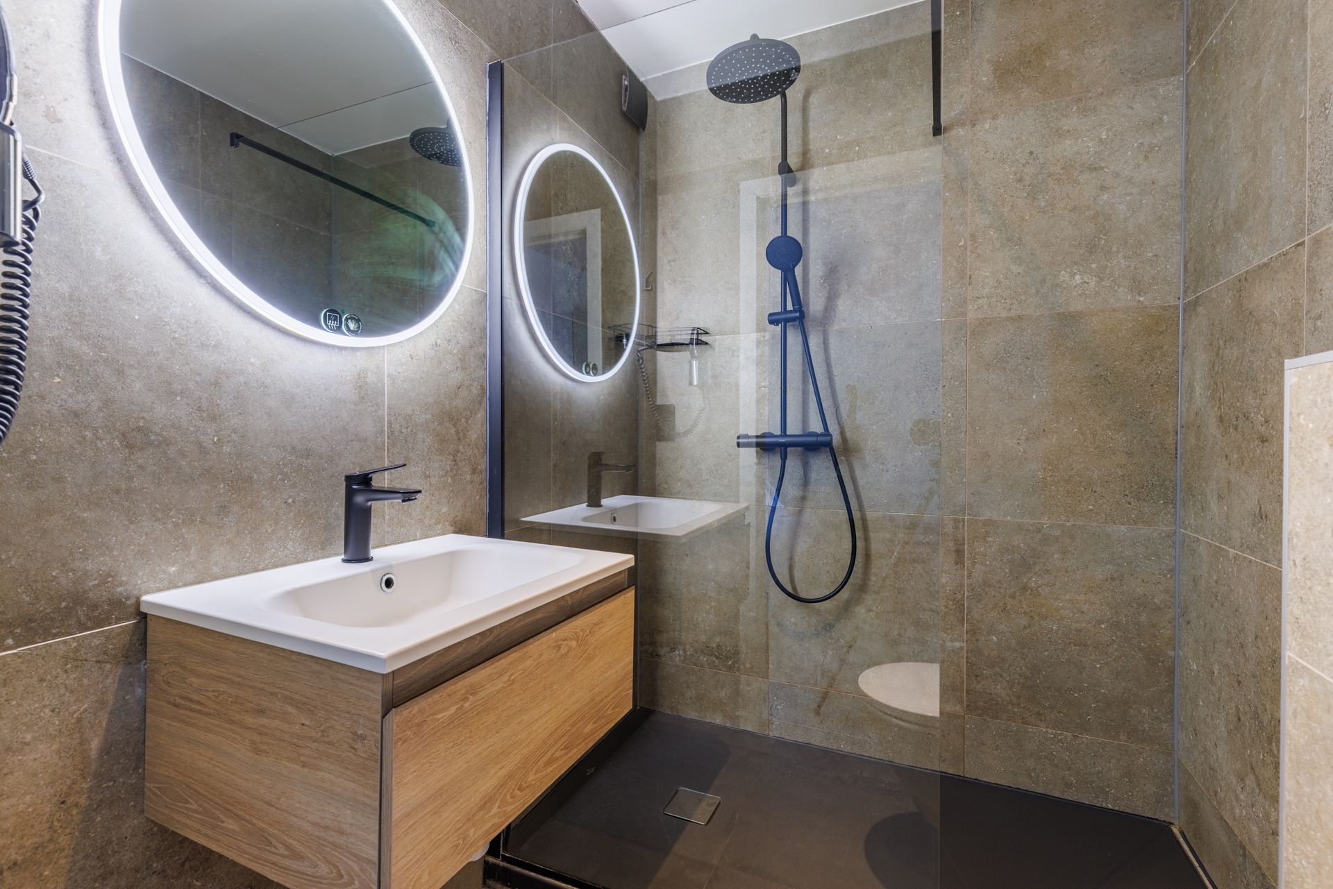 A modern bathroom featuring a floating wooden vanity with a white sink, large backlit circular mirrors, and a glass shower.