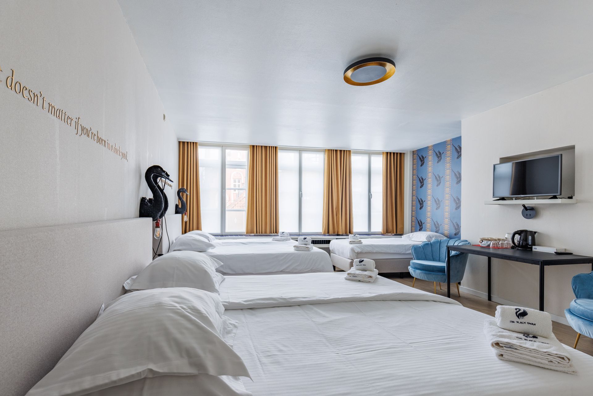 A hotel room featuring three white beds with linens, gold curtains, blue accent wallpaper, a wall-mounted TV, and chairs.