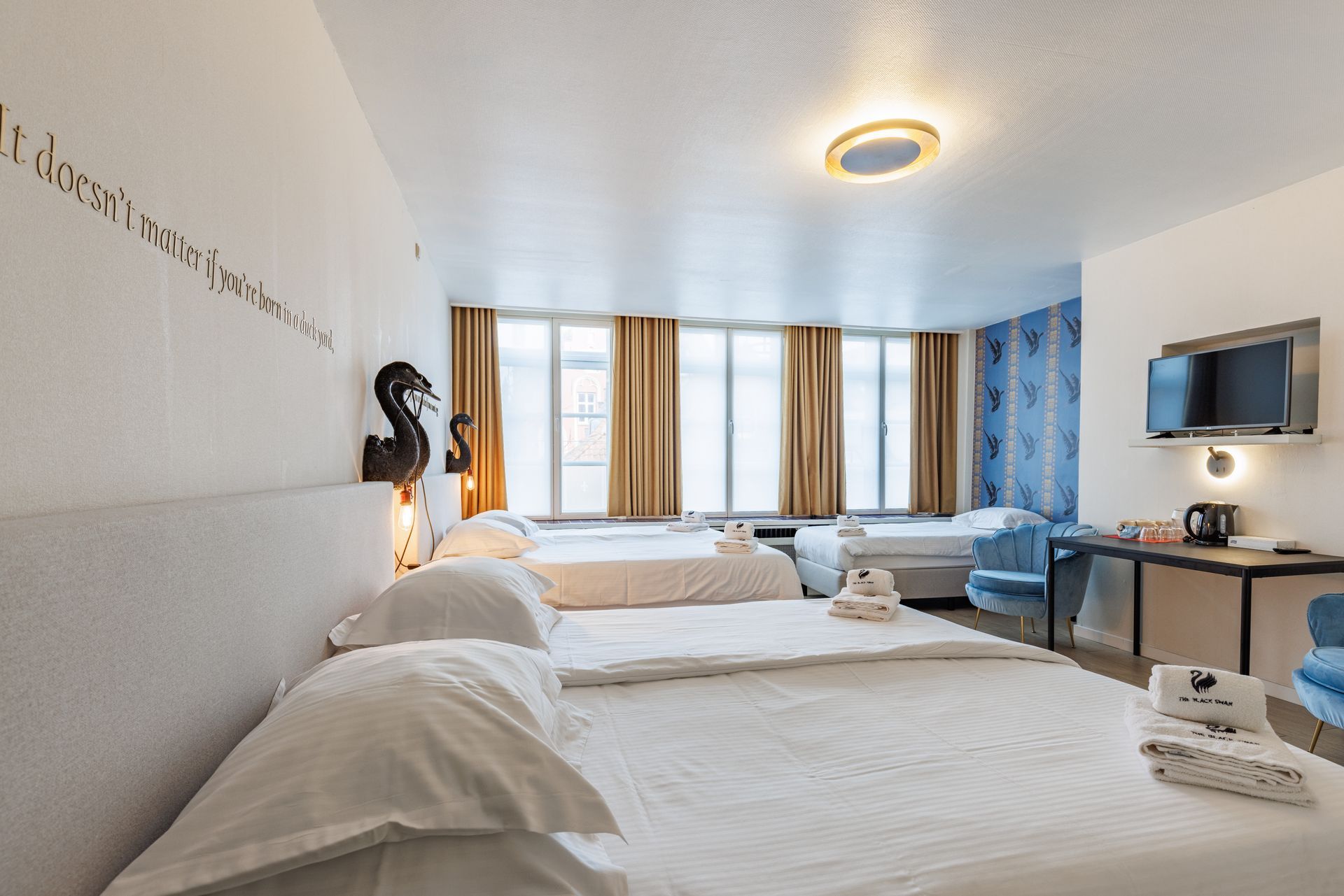 A hotel room with multiple beds, white linens, beige curtains, a television on the wall, and blue accent wallpaper.