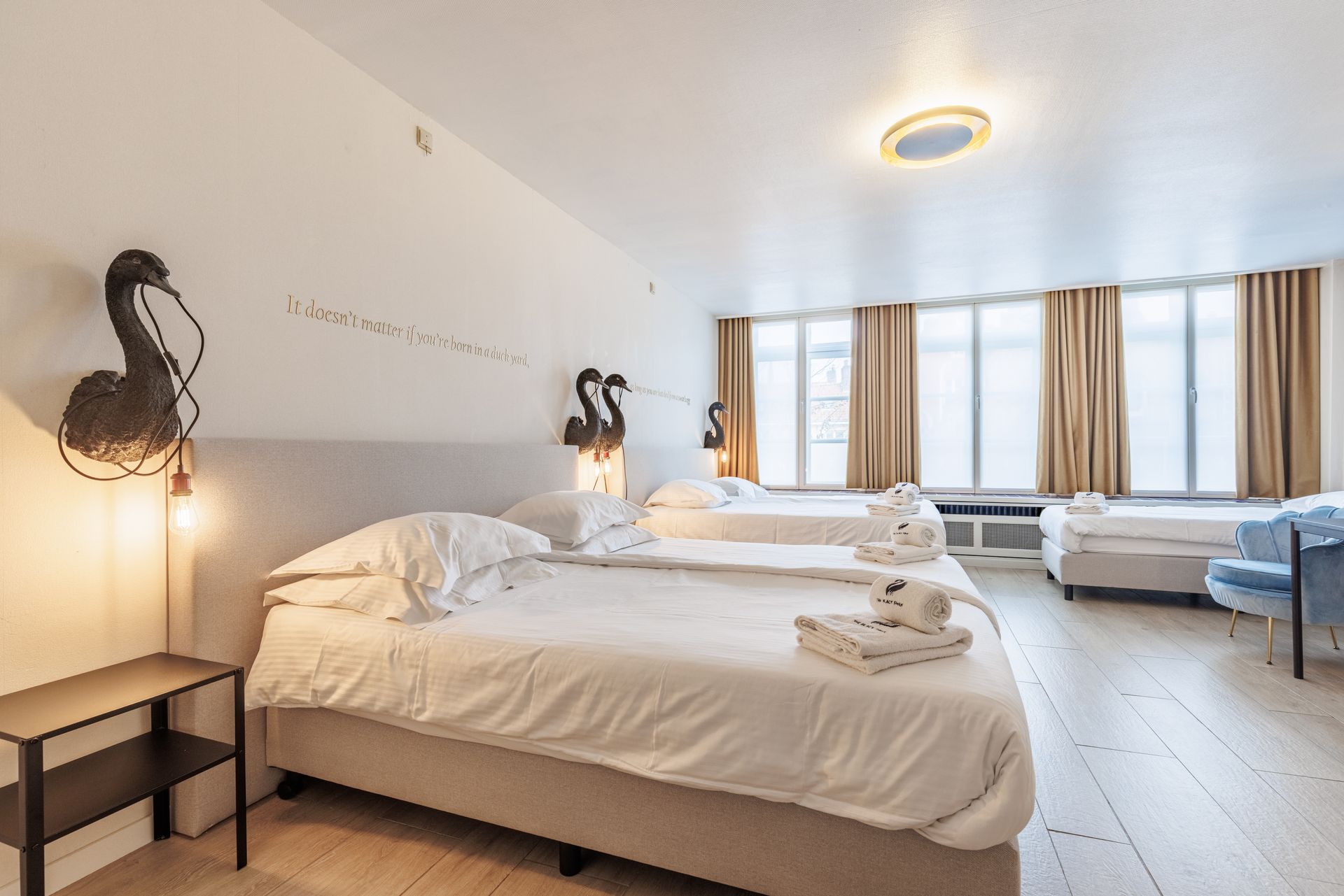 A modern hotel room with three white beds, beige curtains, light wooden floors, and swan-shaped wall sconces.