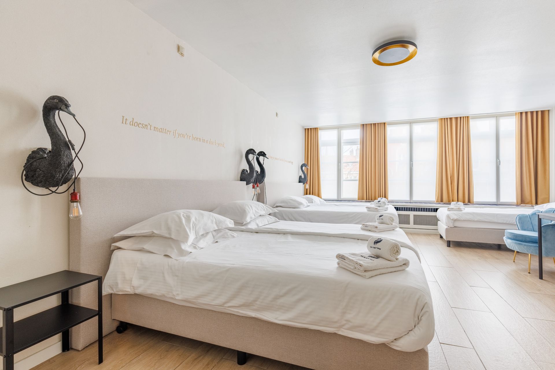 A minimalist hotel room with two white beds, light wood floors, beige curtains, and black swan wall decor.