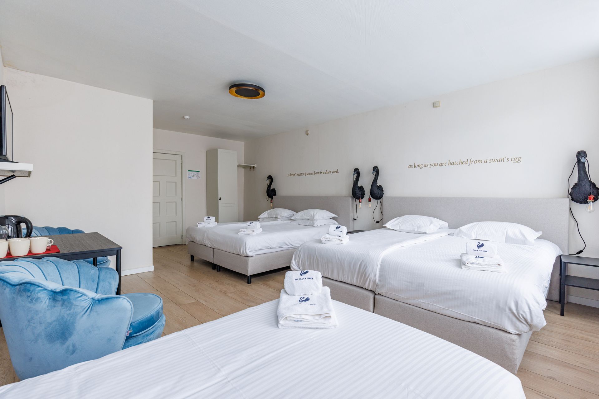 A bright hotel room with three beds featuring white linens, a blue armchair, and flamingo wall decor.