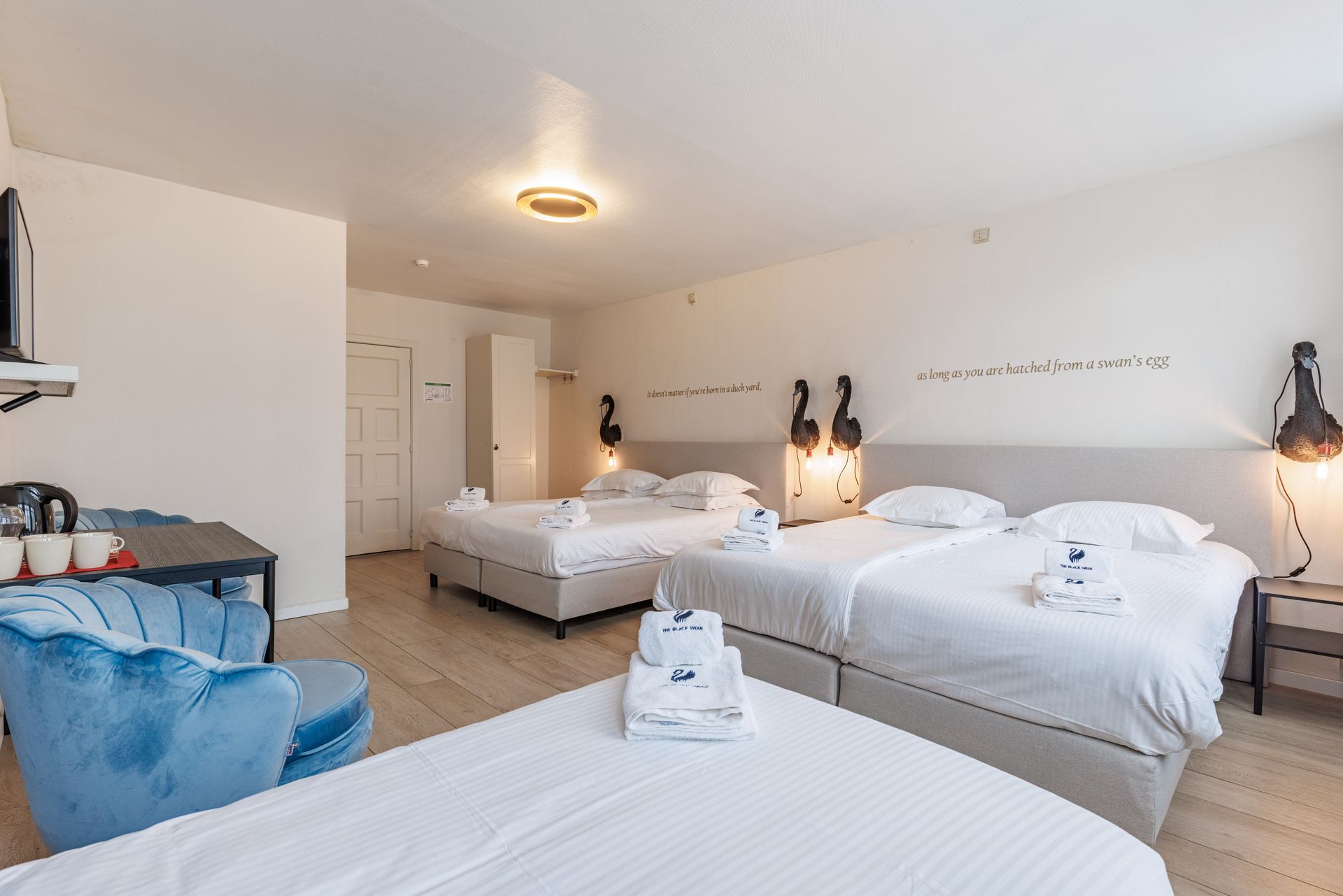 A hotel room featuring three beds with white linens, a blue velvet armchair, and light wood flooring.