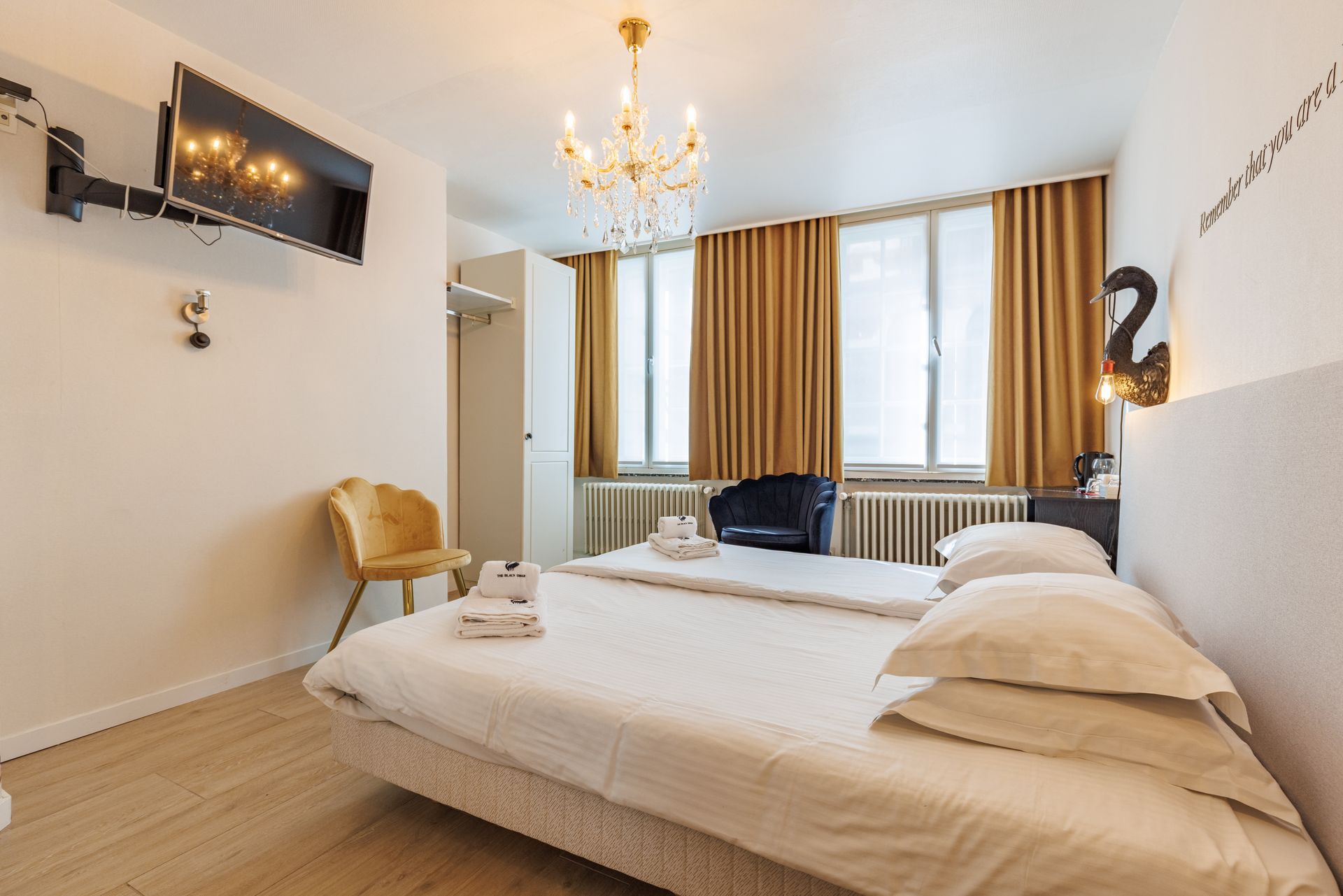 A cozy bedroom featuring a bed with white linens, a crystal chandelier, gold curtains, and a yellow accent chair.