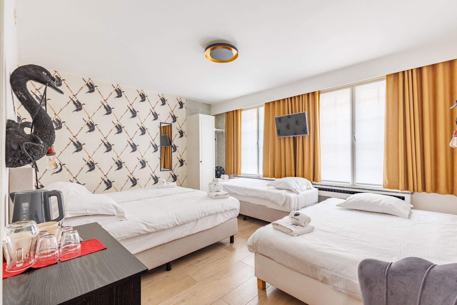 A hotel room with three beds, patterned wallpaper featuring bird silhouettes, a dark side table, and mustard-yellow curtains.