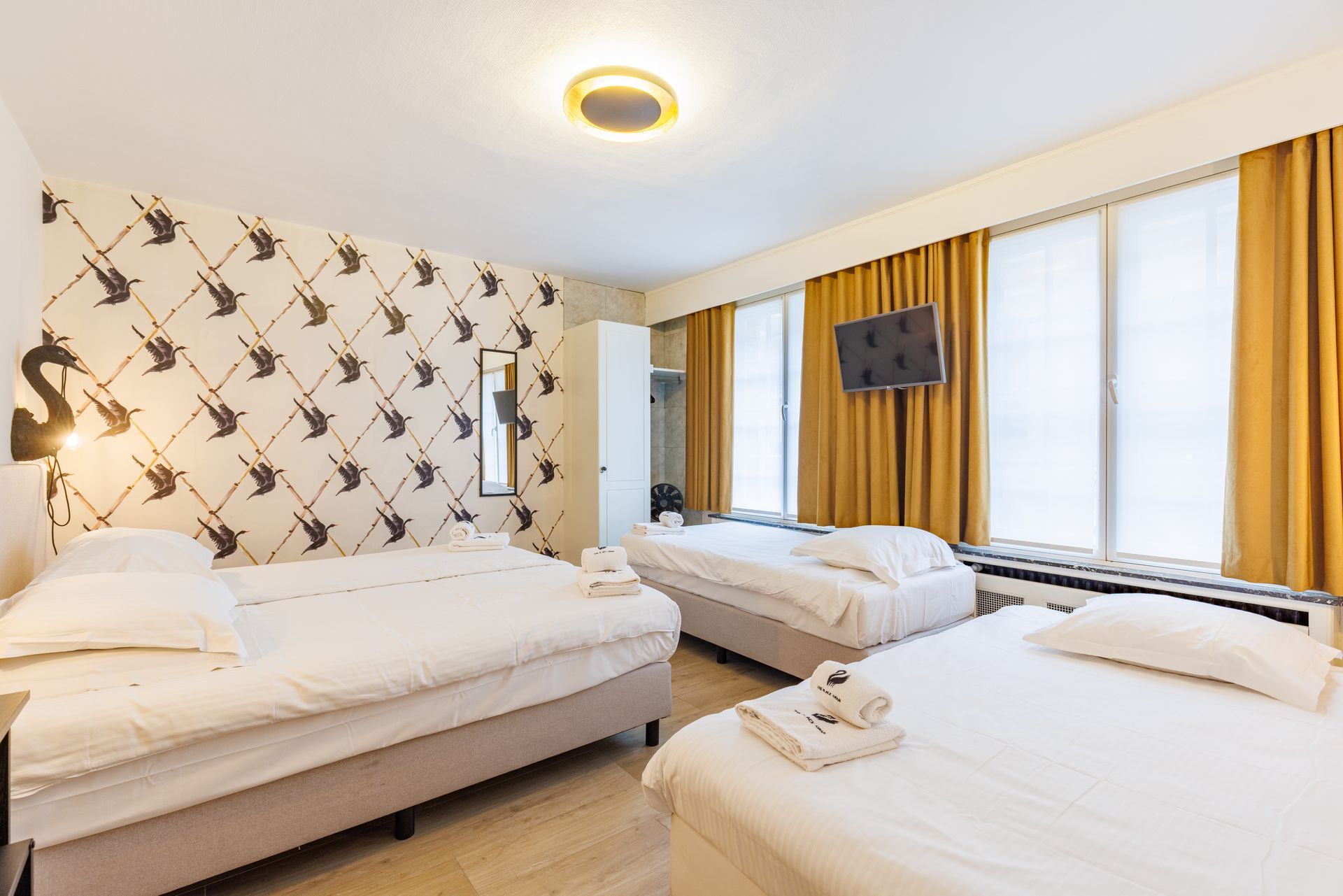 A hotel room featuring three white beds, patterned wallpaper, a wall-mounted TV, and yellow curtains.