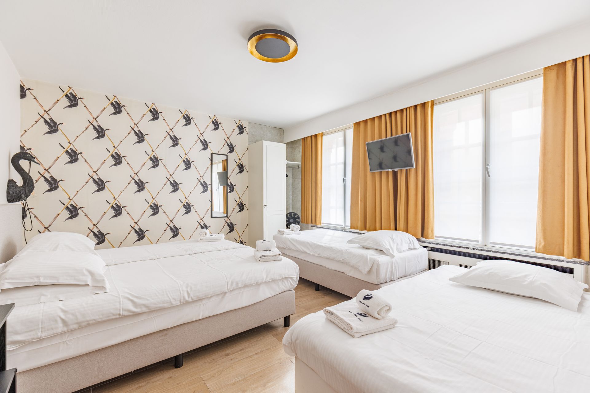 A bright hotel room with three white beds, bird-patterned wallpaper, yellow curtains, and a television on the wall.