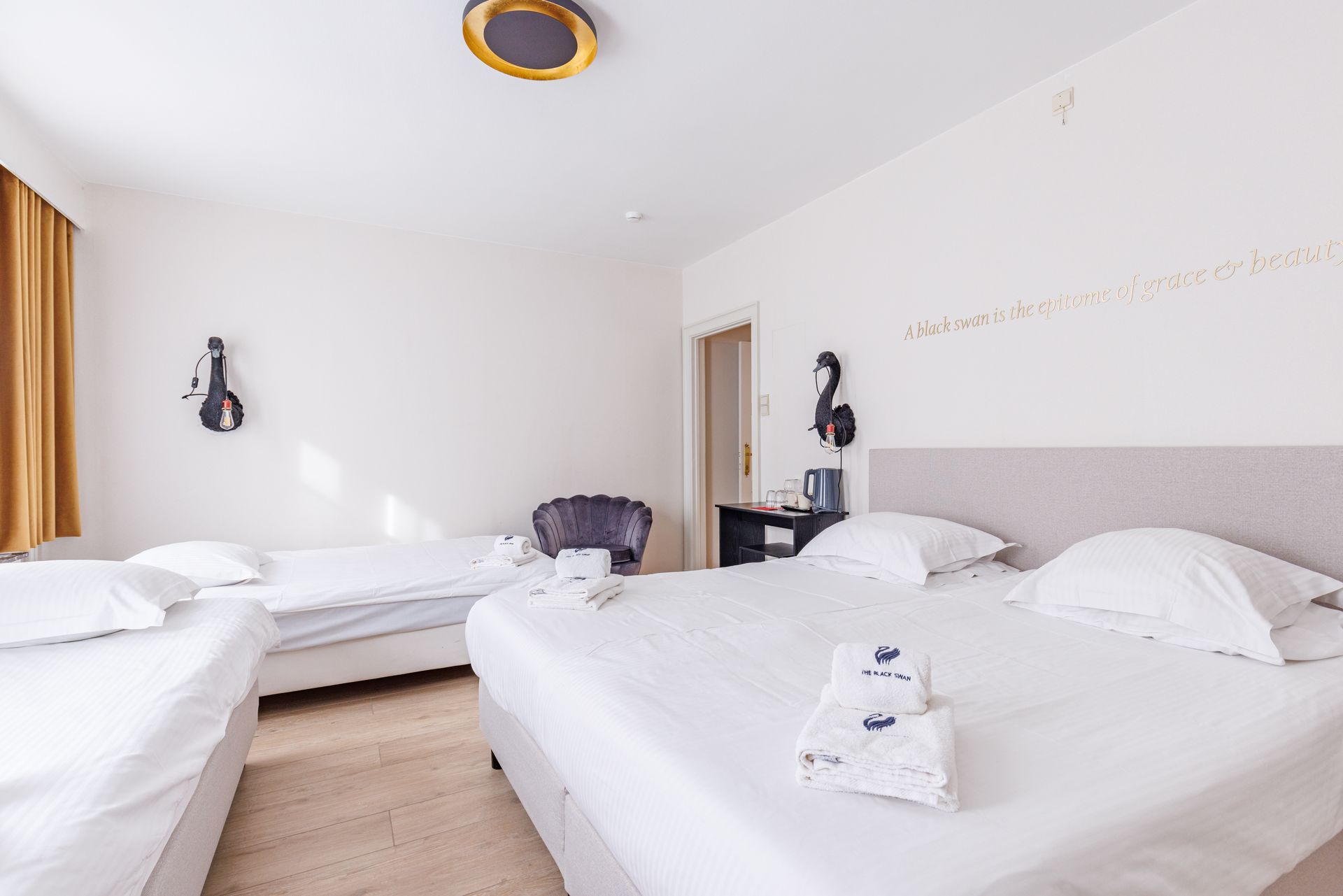 A bright hotel room with a large bed in the foreground and a second bed to the left, featuring white linens and decor.