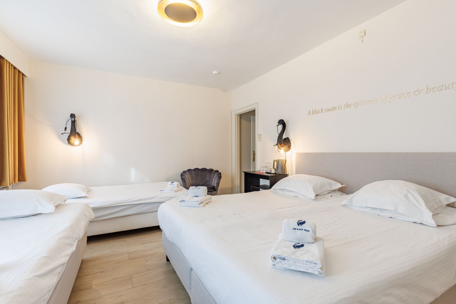 A hotel room with two single beds, one double bed, white bedding, wall sconces, a small chair, and a light wooden floor.