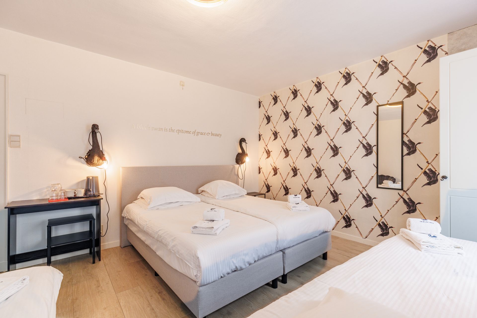 A hotel room with two twin beds side-by-side, light wood floors, a desk, and a patterned wallpaper featuring birds.