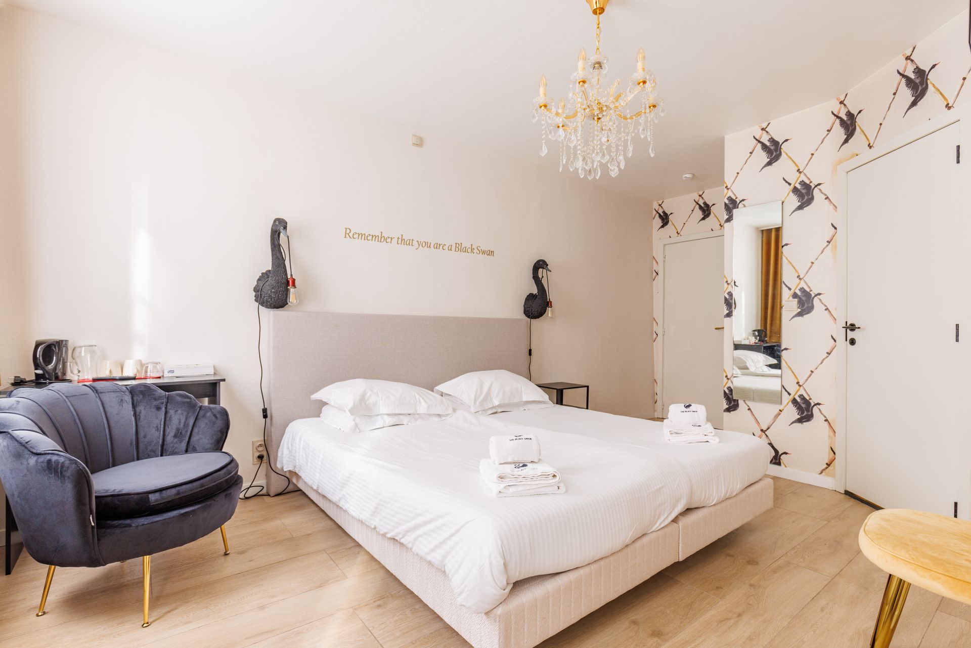 A brightly lit hotel bedroom with a large bed, grey armchair, gold chandelier, and bird-patterned wallpaper.