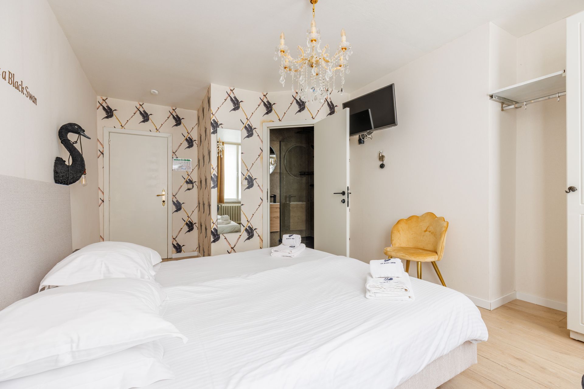 A bright hotel room features a large bed with white linens, a crystal chandelier, patterned wallpaper, and a gold chair.