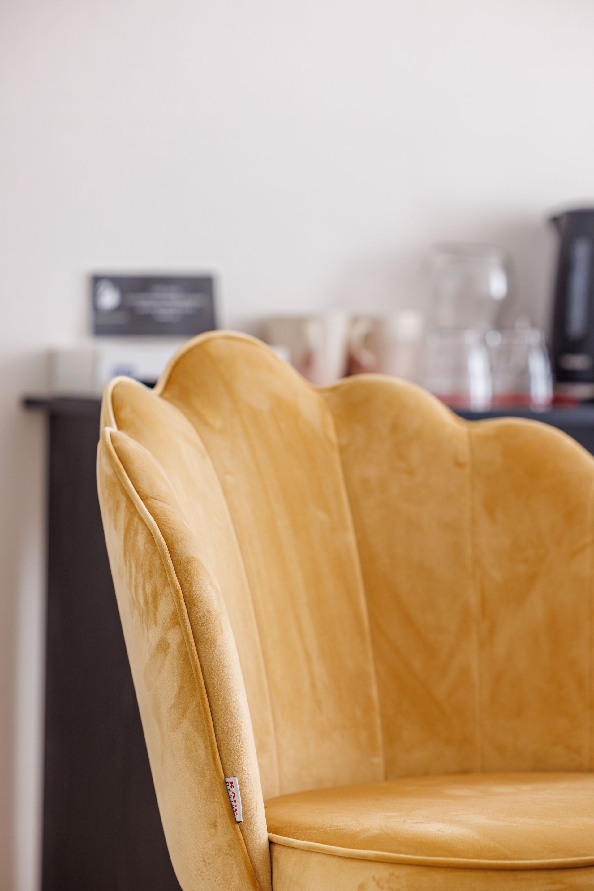A close-up of a mustard-yellow velvet chair with a scalloped, shell-shaped back in a blurred interior setting.