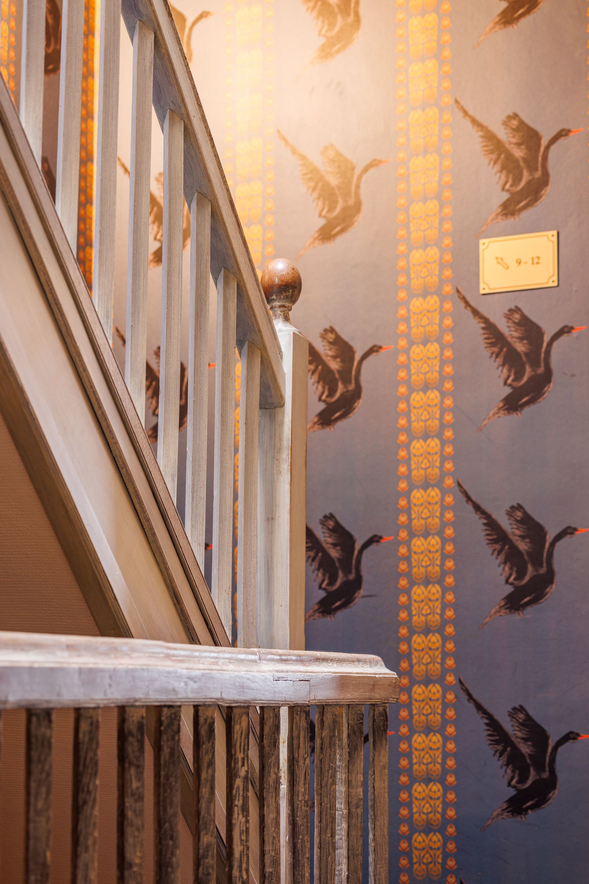 A wooden staircase with pale railings set against blue wallpaper featuring repeating patterns of birds and gold columns.