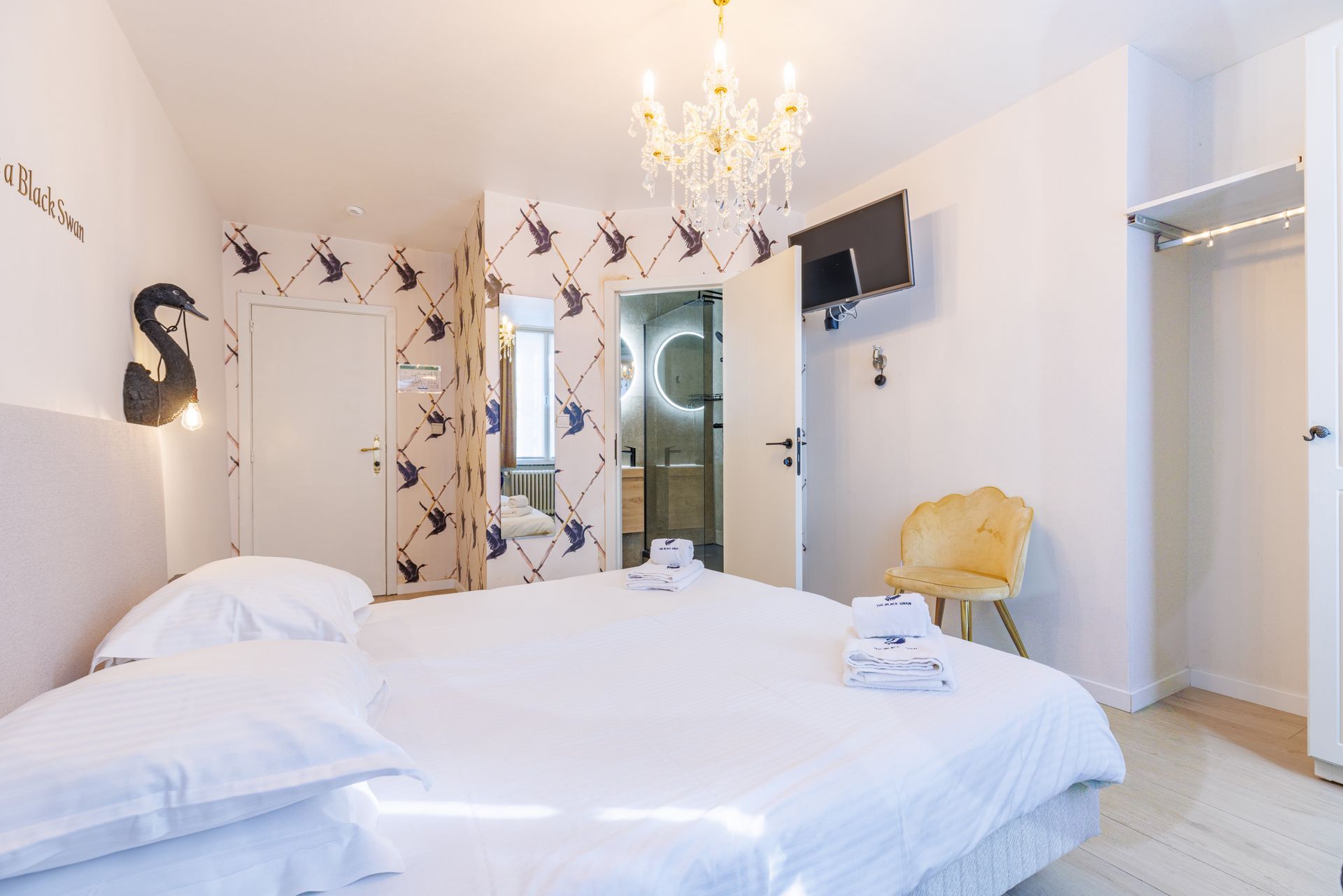 A bright hotel bedroom with a white bed, floral wallpaper, a crystal chandelier, and a yellow accent chair.