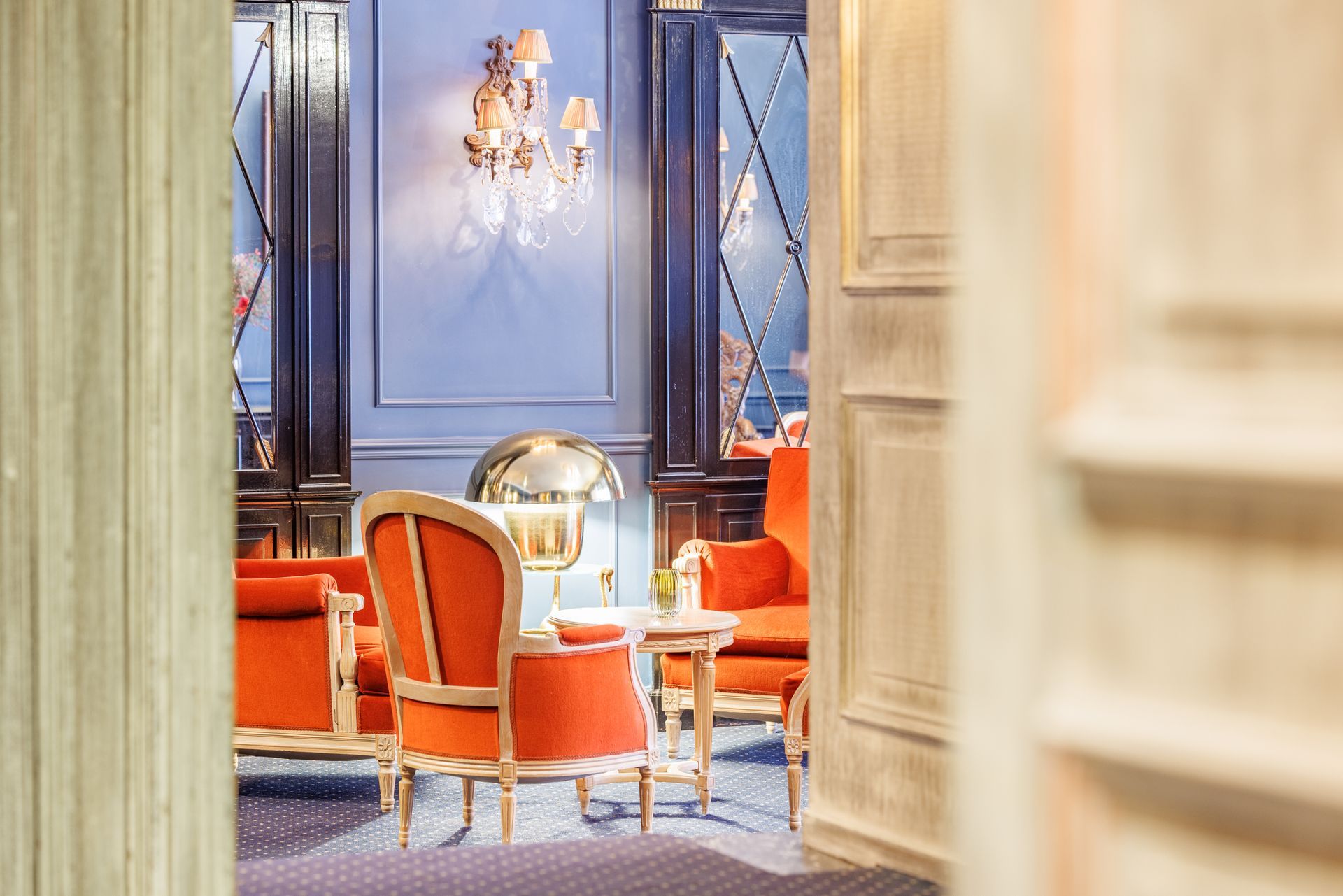 A pair of orange armchairs and a small side table arranged in a blue room with ornate mirrors and a wall sconce.