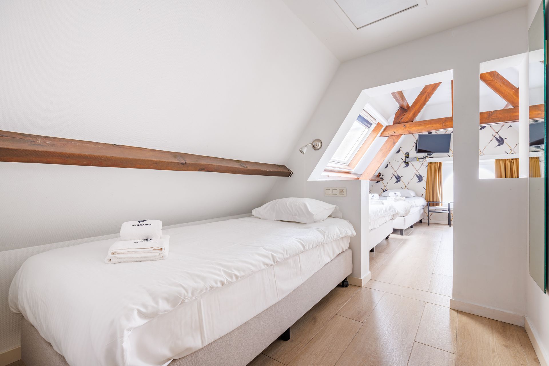 Attic bedroom with a white single bed, wooden beams, and an adjacent sleeping area seen through an opening.