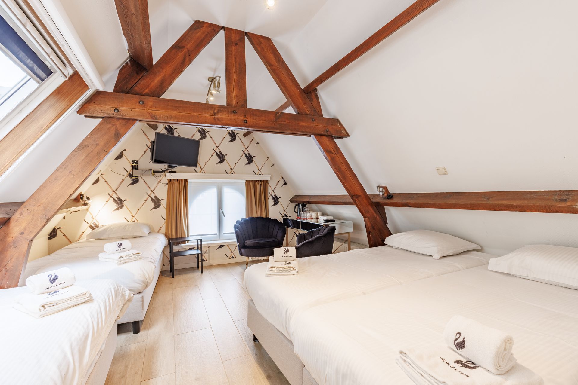 An attic hotel room featuring wooden beams, slanted ceilings, three made beds, and a small sitting area by a window.