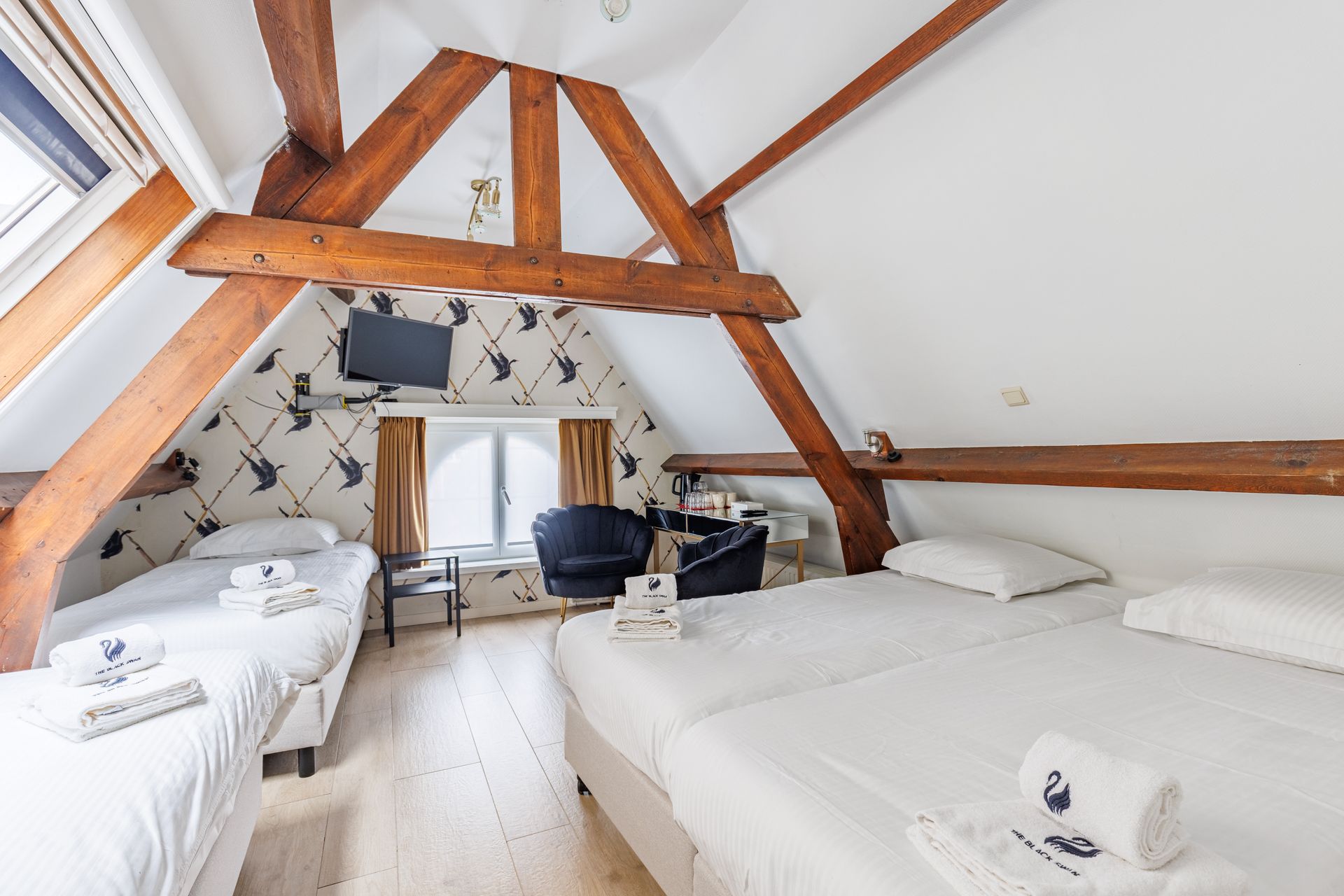 Attic bedroom with three twin beds, wooden beams, sloped ceilings, and wallpaper featuring black swan silhouettes.
