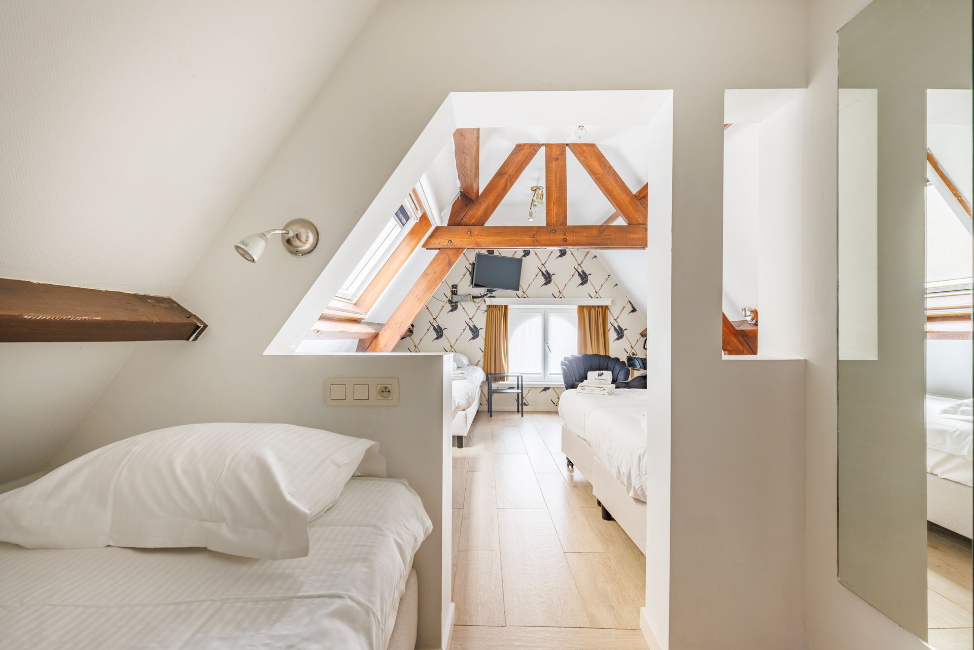A bright attic hotel suite with a single bed in the foreground and a larger bedroom area visible through a partition.