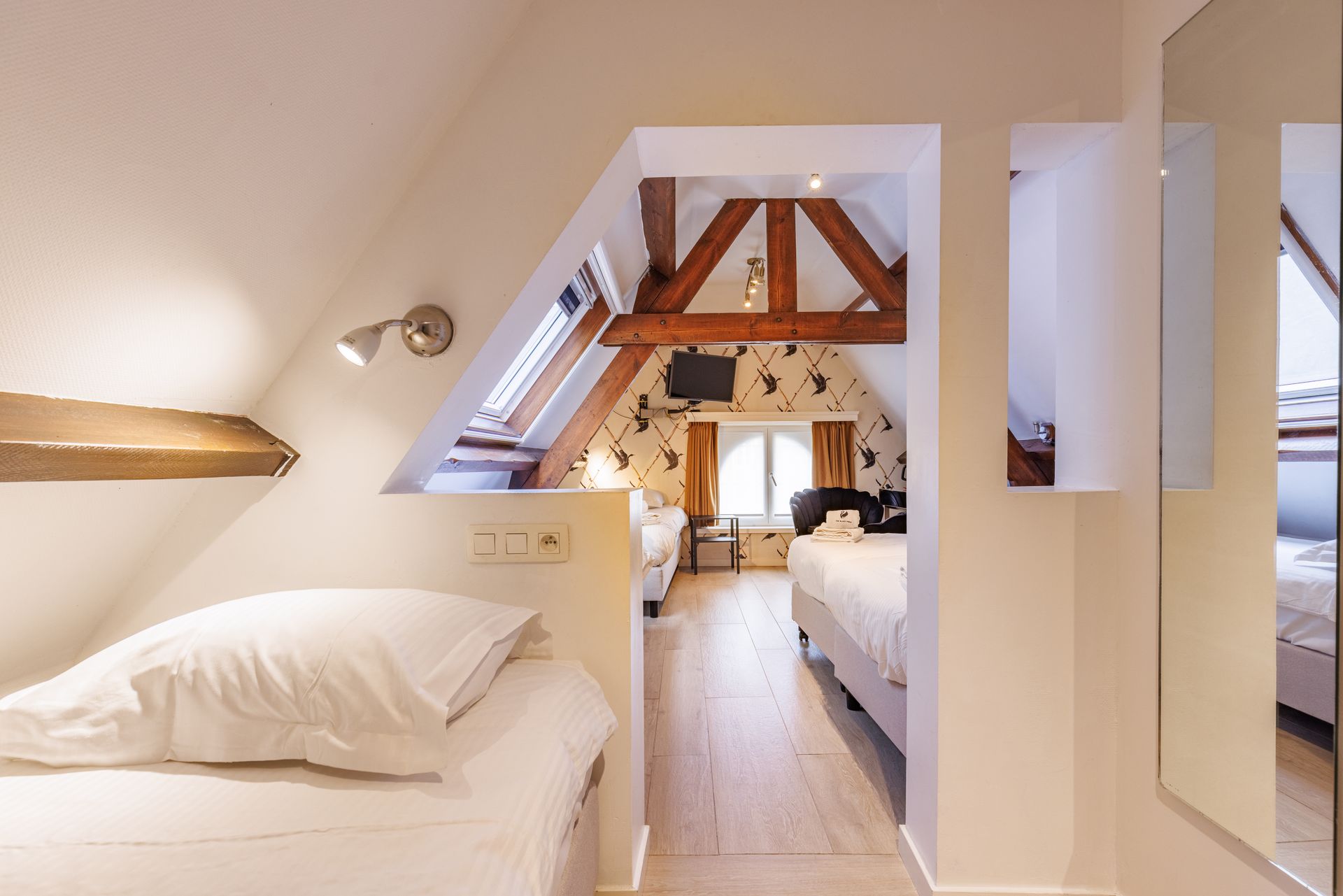 A cozy attic hotel room with a slanted ceiling, wooden beams, and two separate bed areas separated by a partition.