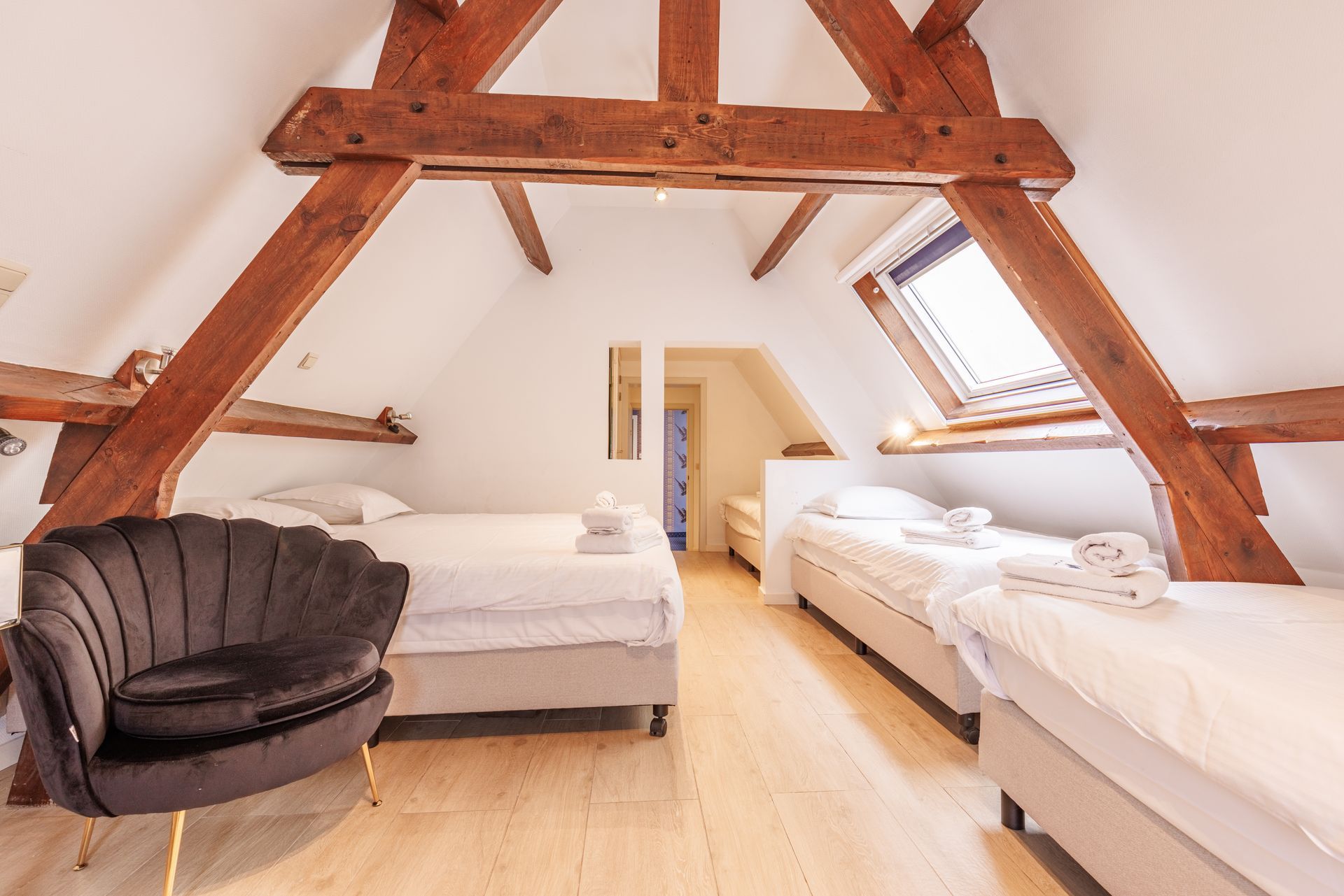Attic bedroom with exposed wooden beams, a charcoal velvet chair, and three white beds under a sloped ceiling and skylight.