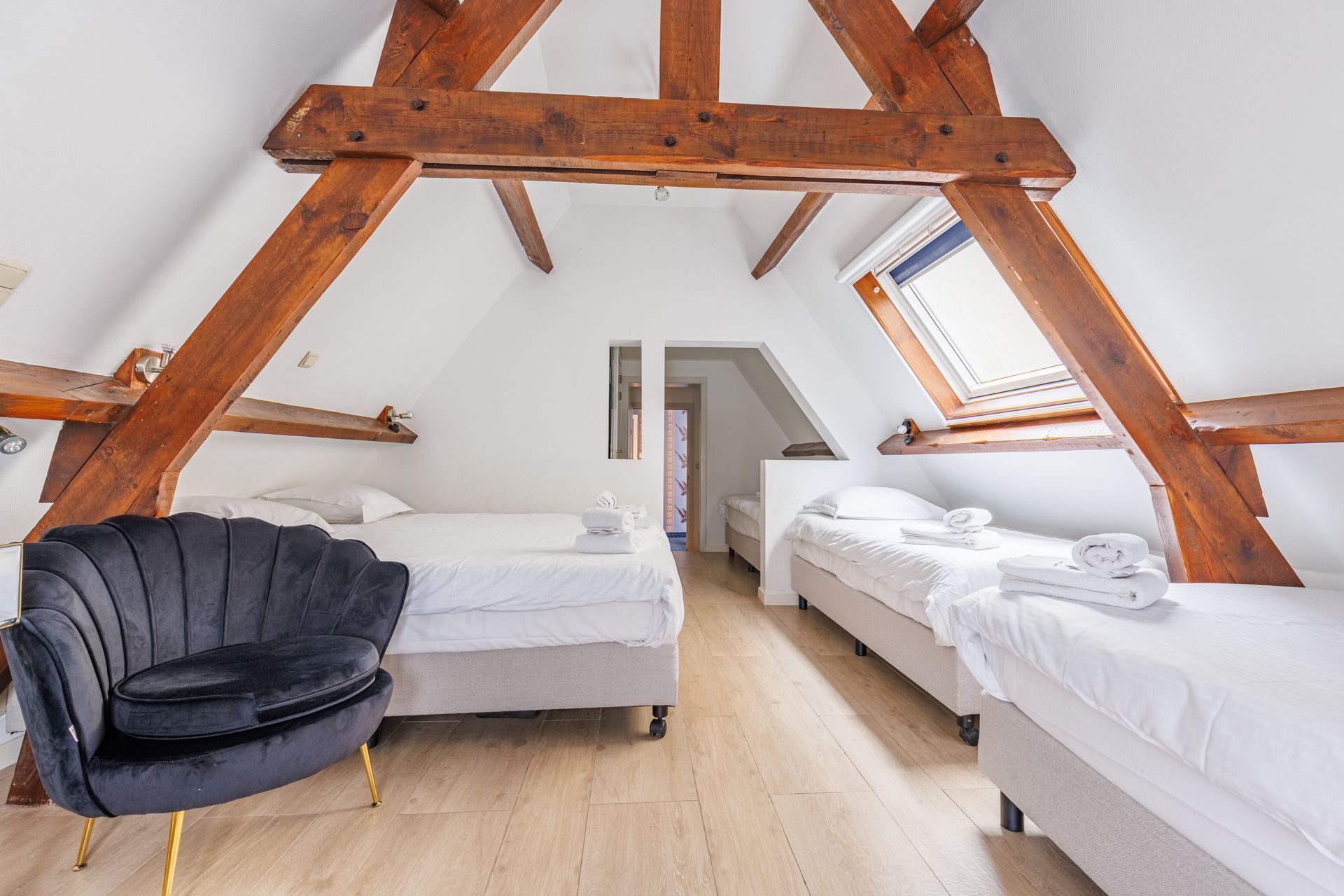An attic bedroom with wooden beams, three beds with white linens, and a black, shell-shaped velvet armchair.